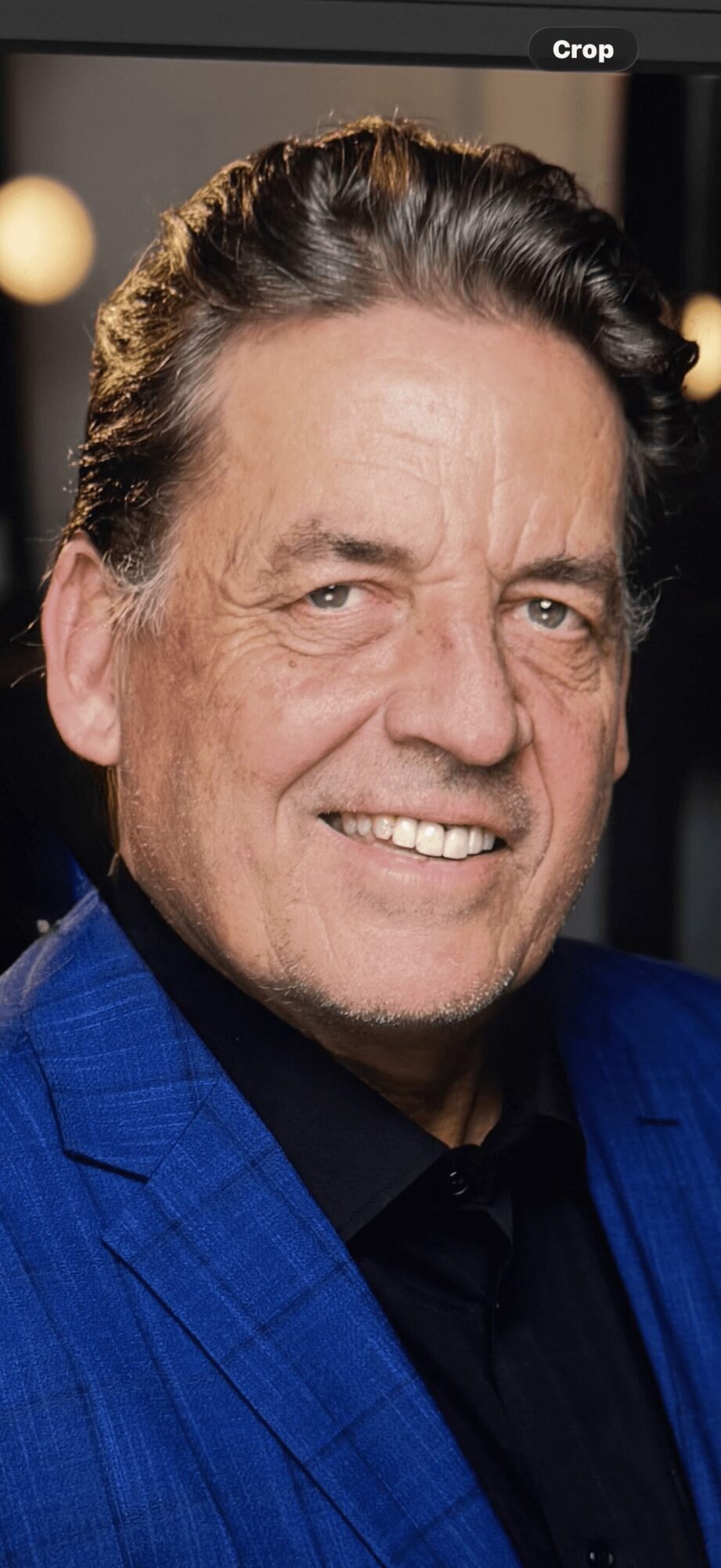 Close-up of a smiling man with dark hair, wearing a blue suit and black shirt.