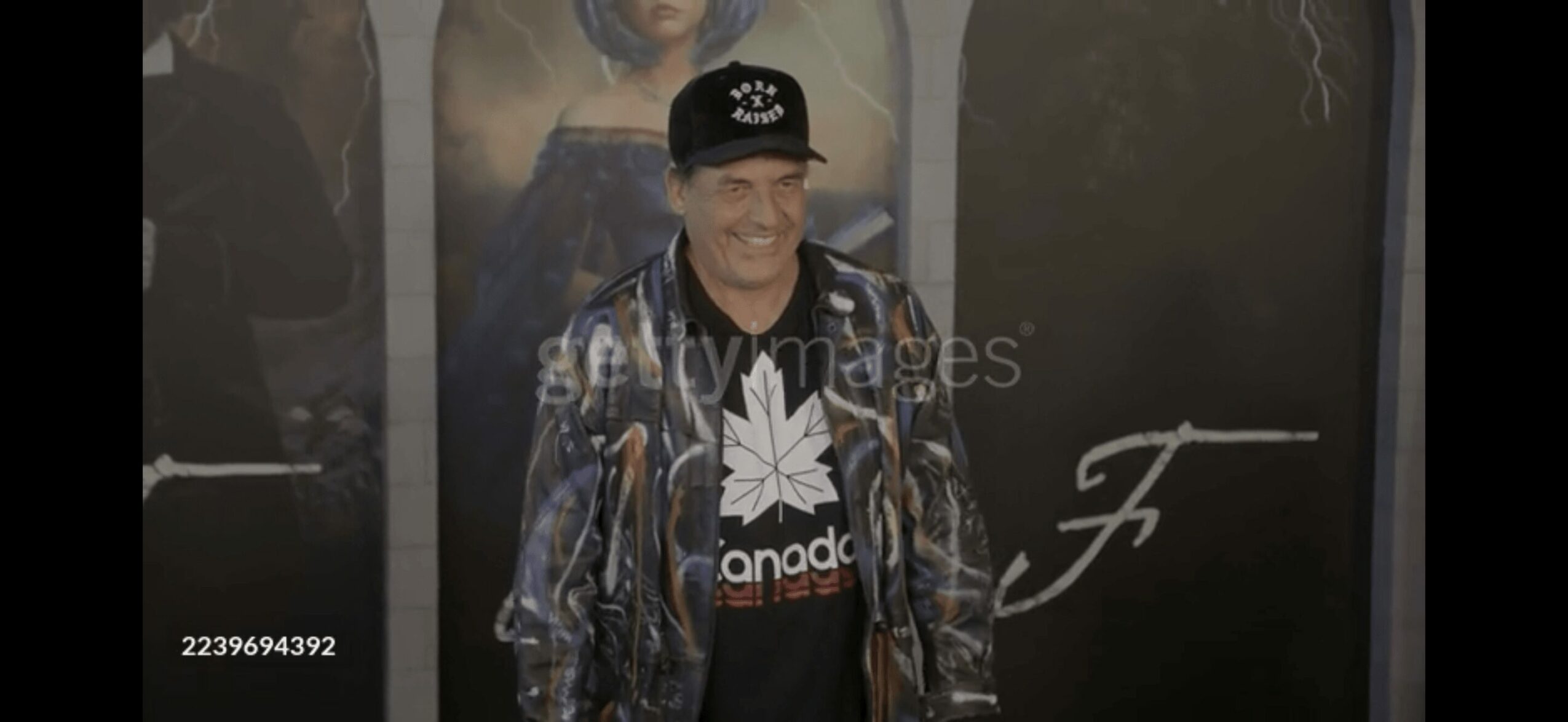 Person wearing a cap and jacket standing in front of a dark background with writing.