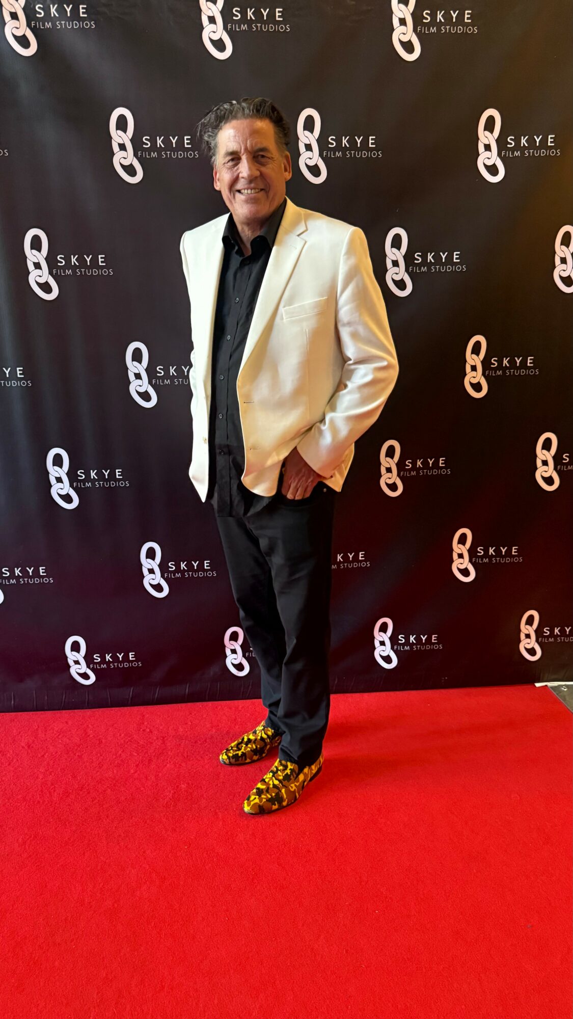 Man in white blazer and black pants standing on red carpet with black backdrop featuring white and orange logo patterns.