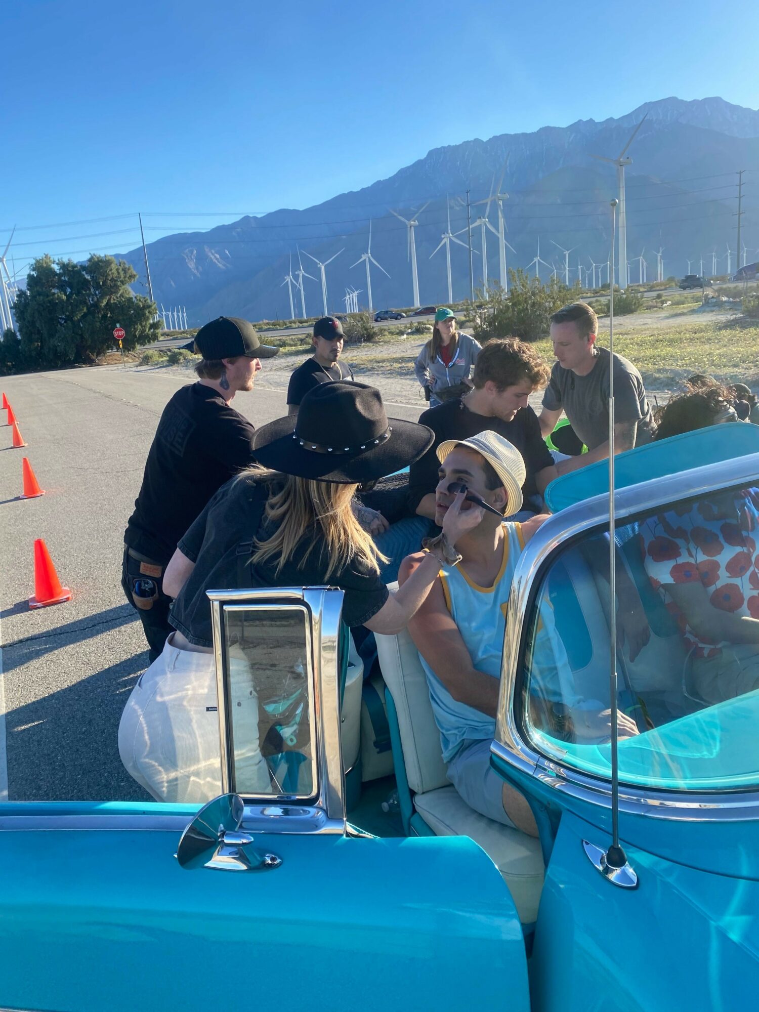 Group of people gathered outdoors near a blue vehicle with mountains and wind turbines in background.