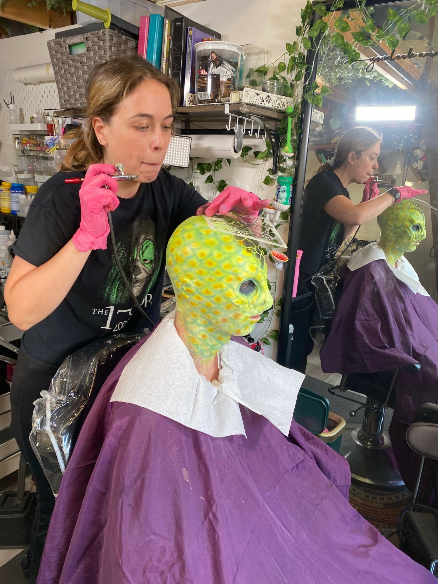 Woman applying green face paint to a person with a yellow-green mask in a salon.