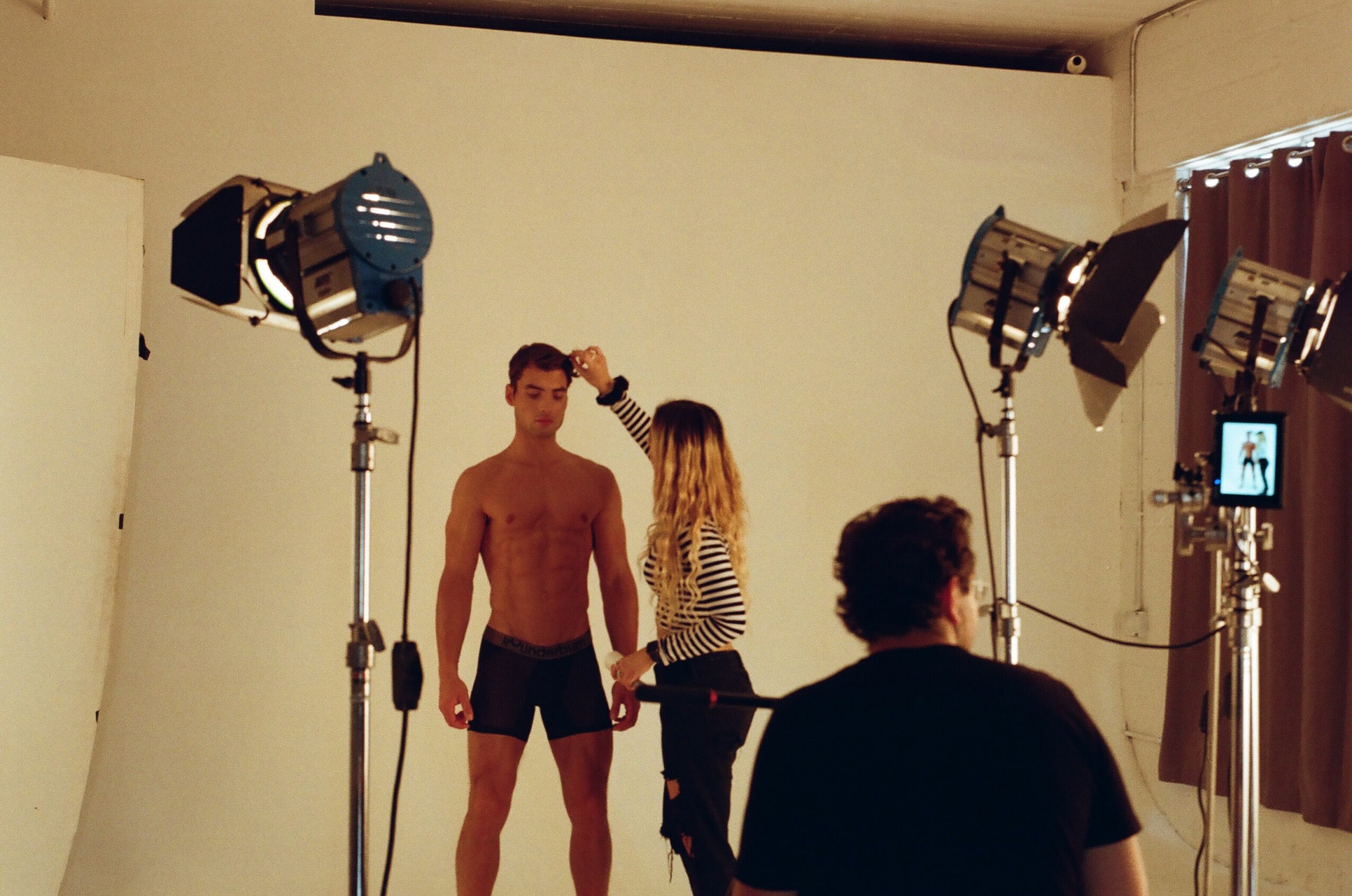 A shirtless man stands in a photography studio with a woman adjusting his hair, surrounded by studio lights and equipment.