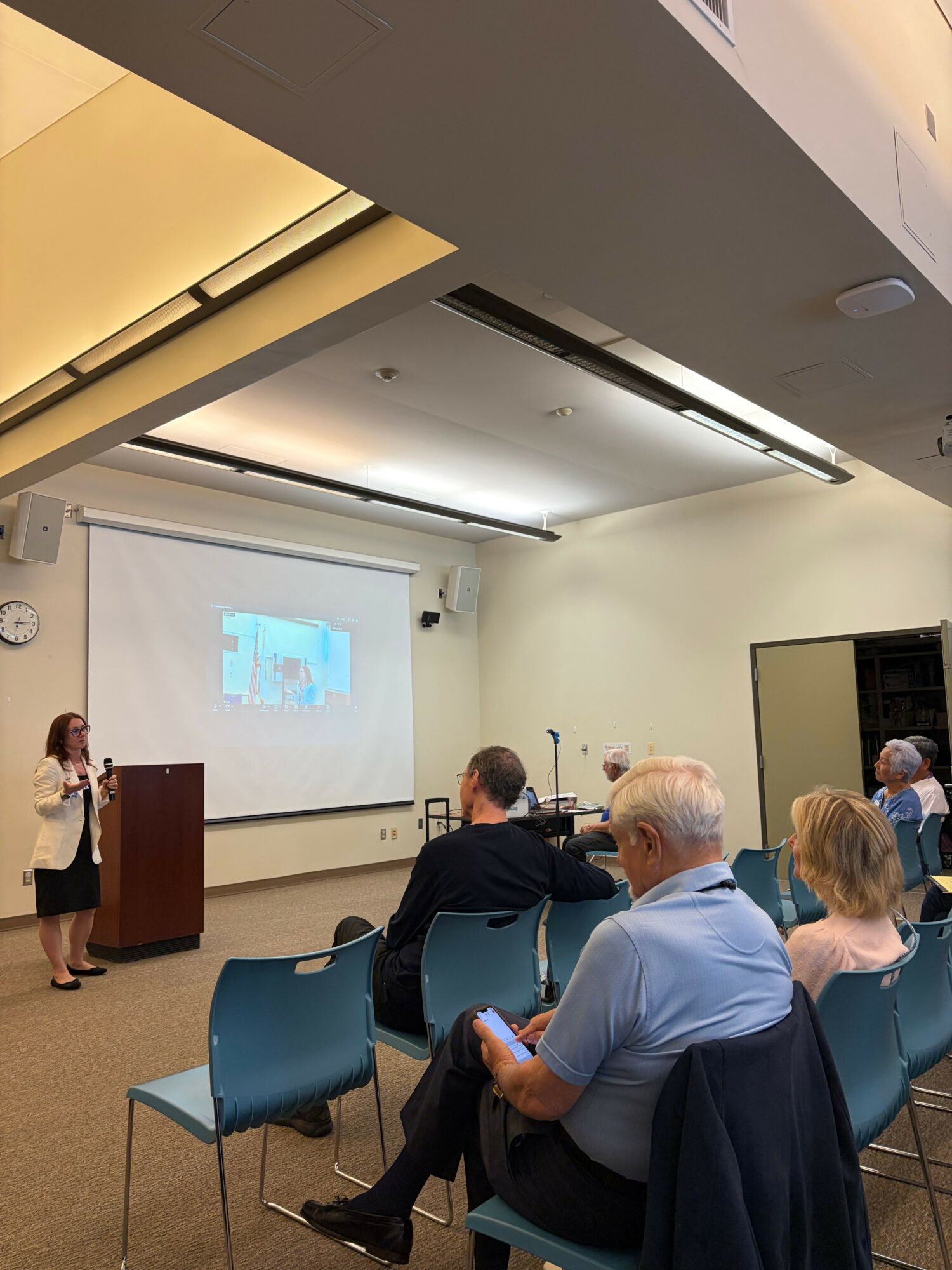A woman presents in front of a projection screen to an audience seated in a conference room.