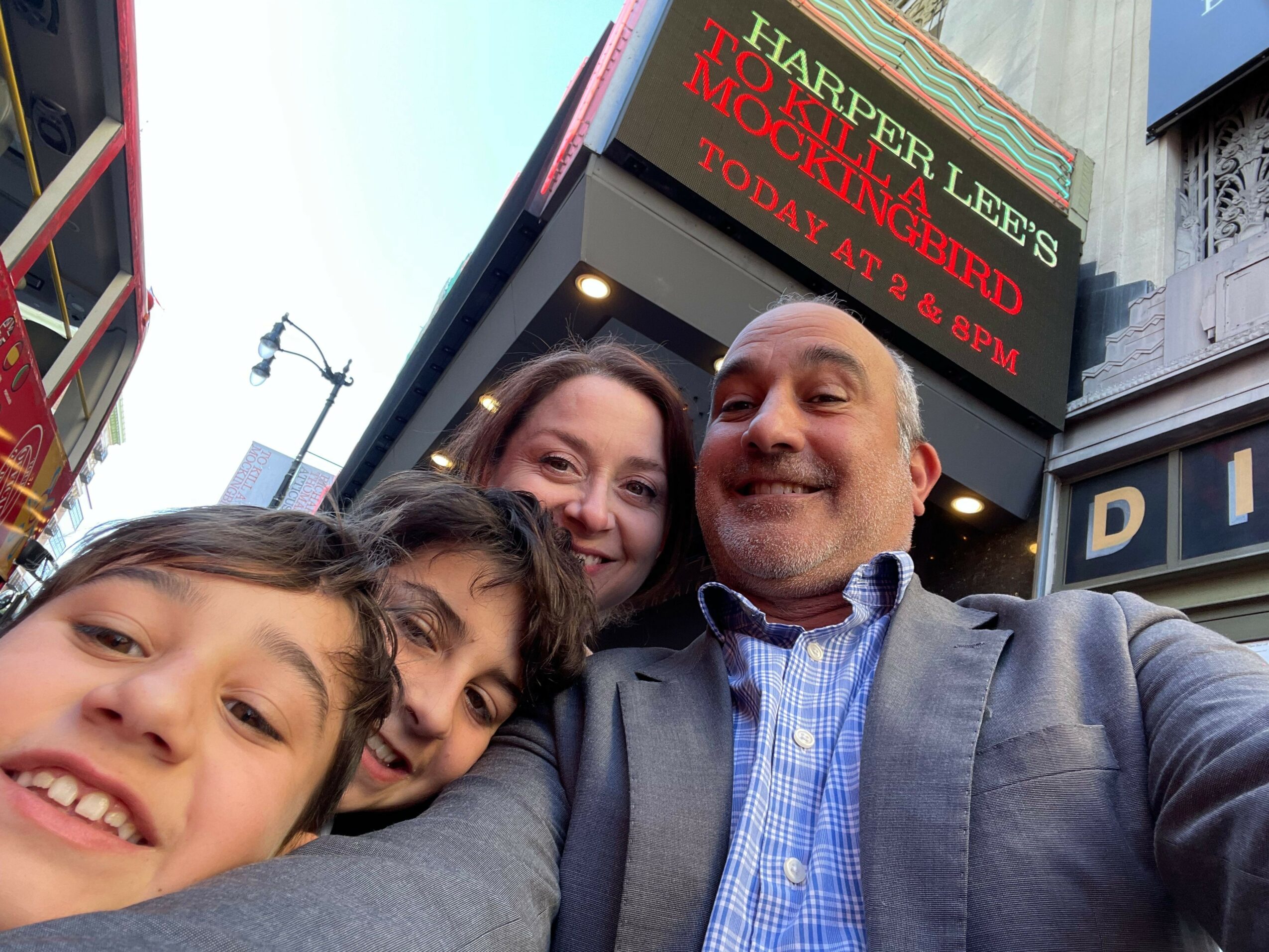 Four people taking a selfie in front of a theater marquee with a sign for Harper Lee's 'To Kill a Mockingbird'.