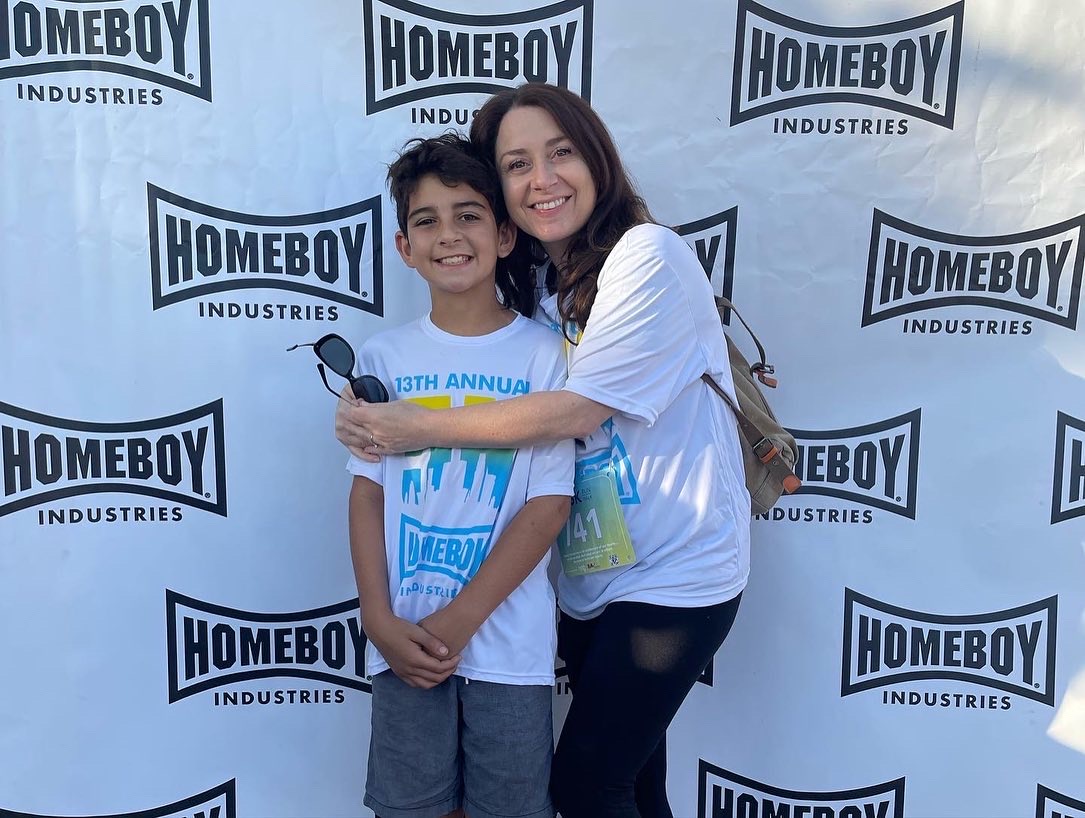 Two people smiling and hugging in front of a backdrop with 'Homeboy Industries' logos.