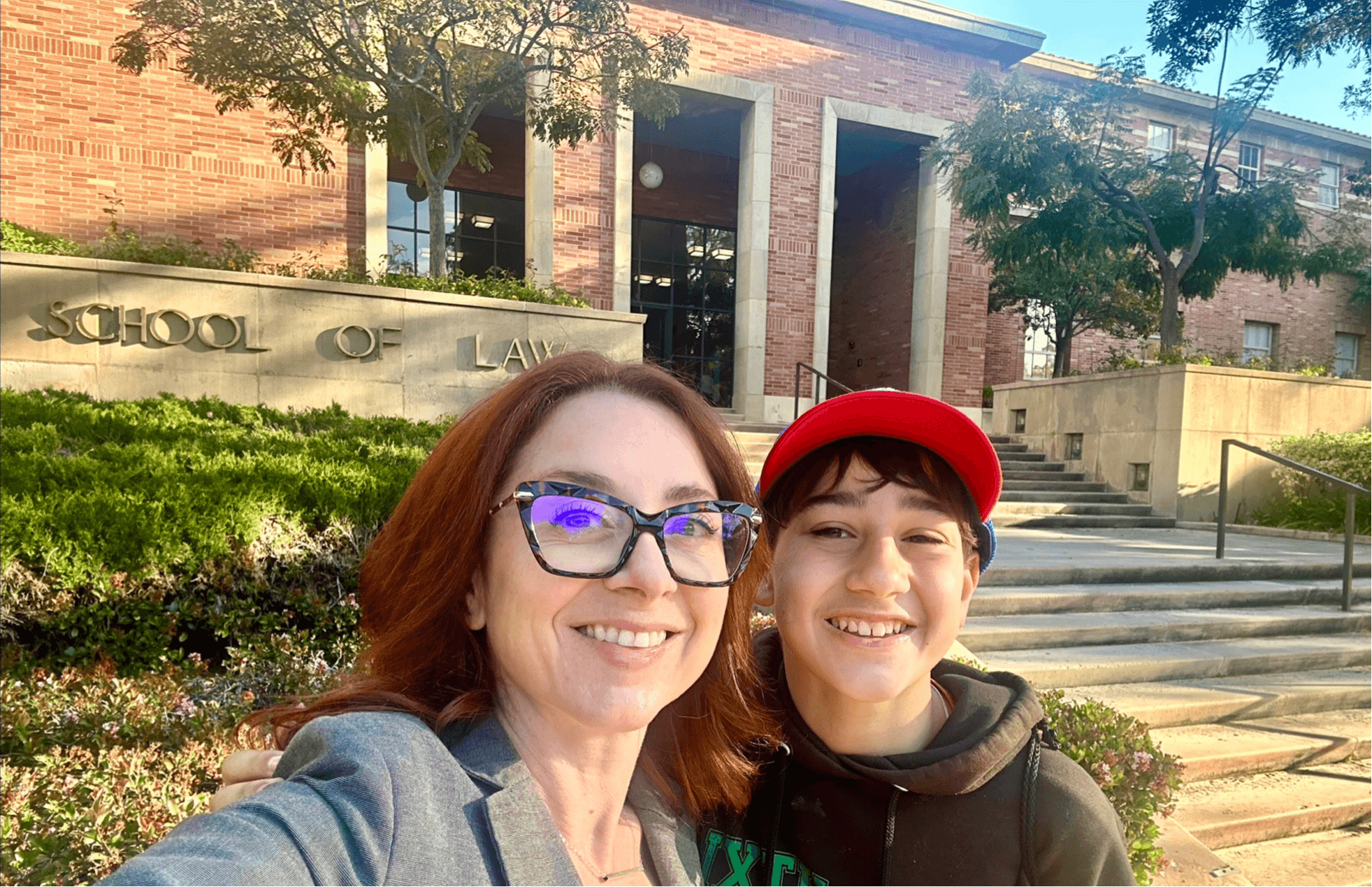 Two people smiling outdoors in front of a building with stairs and trees.