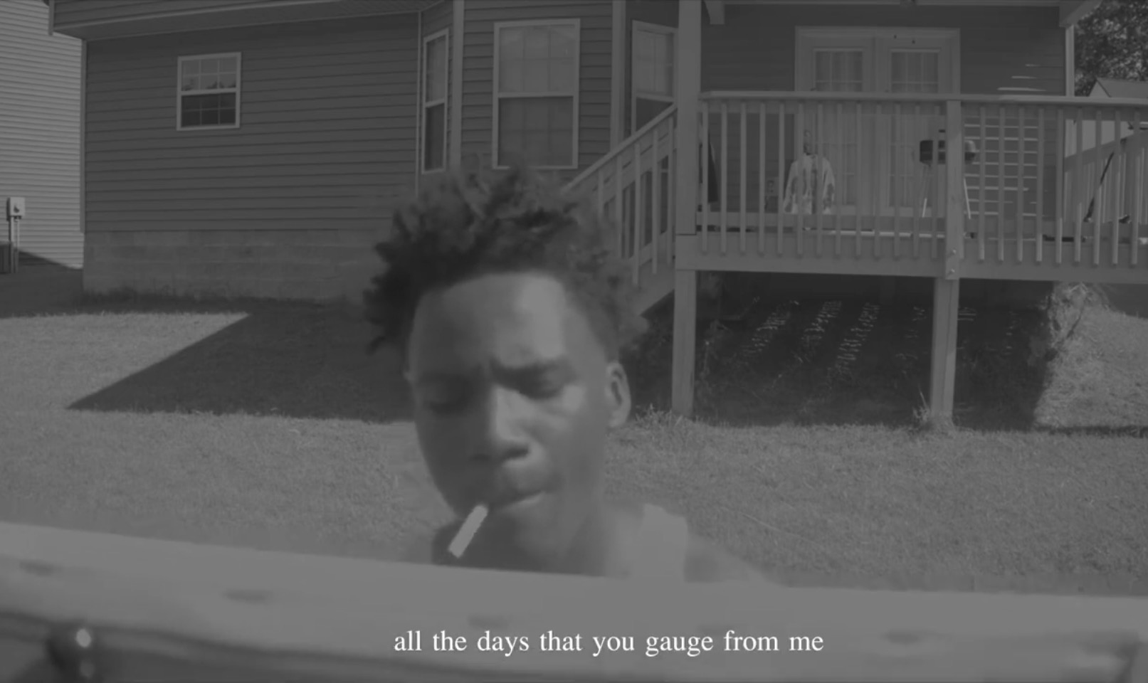 Young person with short hair smoking outdoors in front of a house with a deck.