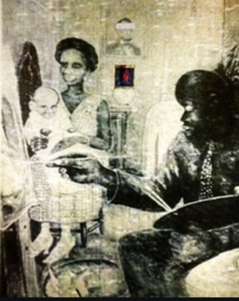 A woman holding a child and a man sitting and pointing at something in a room with a lamp and wall decorations.