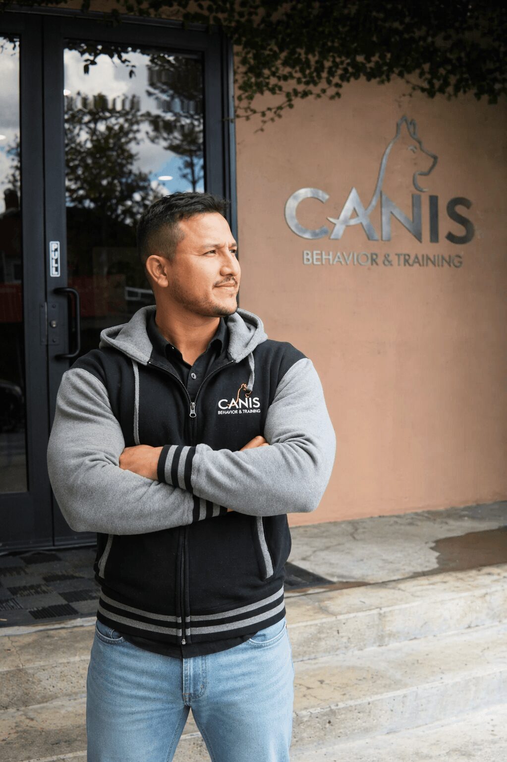 Man standing outside building with arms crossed, wearing a black and gray jacket, in front of a sign that reads 'CANIS Behavior & Training'.