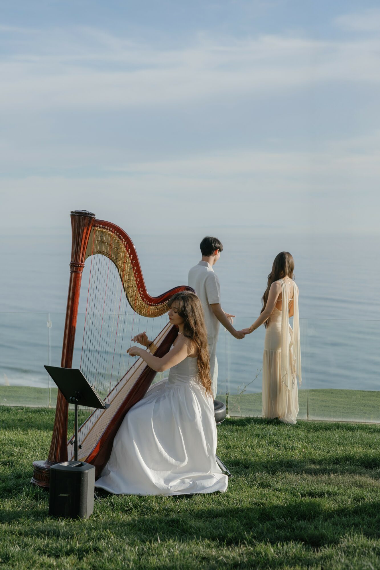 Two women in white dresses and a man in a white shirt by the water, one woman playing a harp, others holding hands.