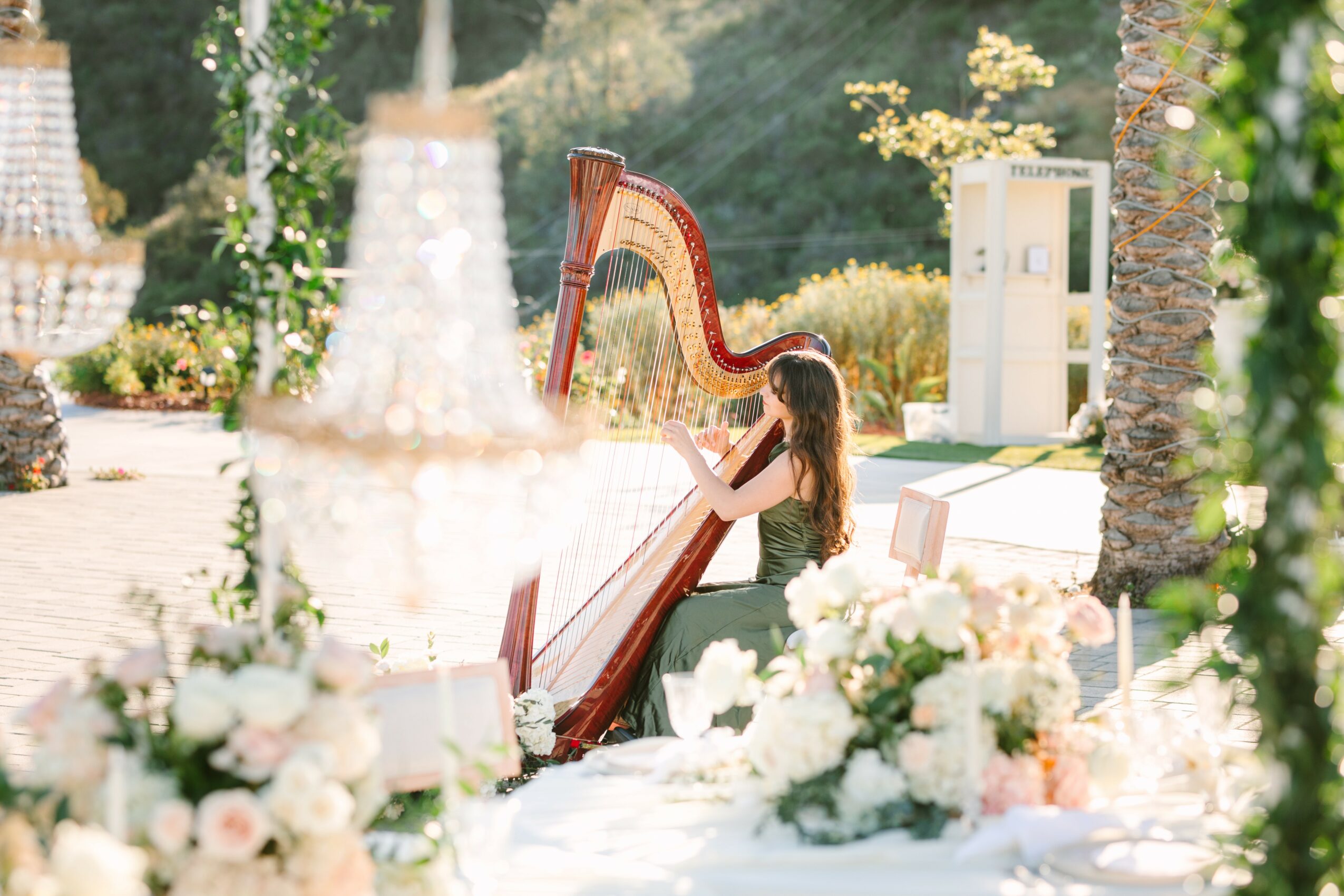 Woman playing harp outdoors surrounded by flowers and greenery, with tables and decorations in the foreground.