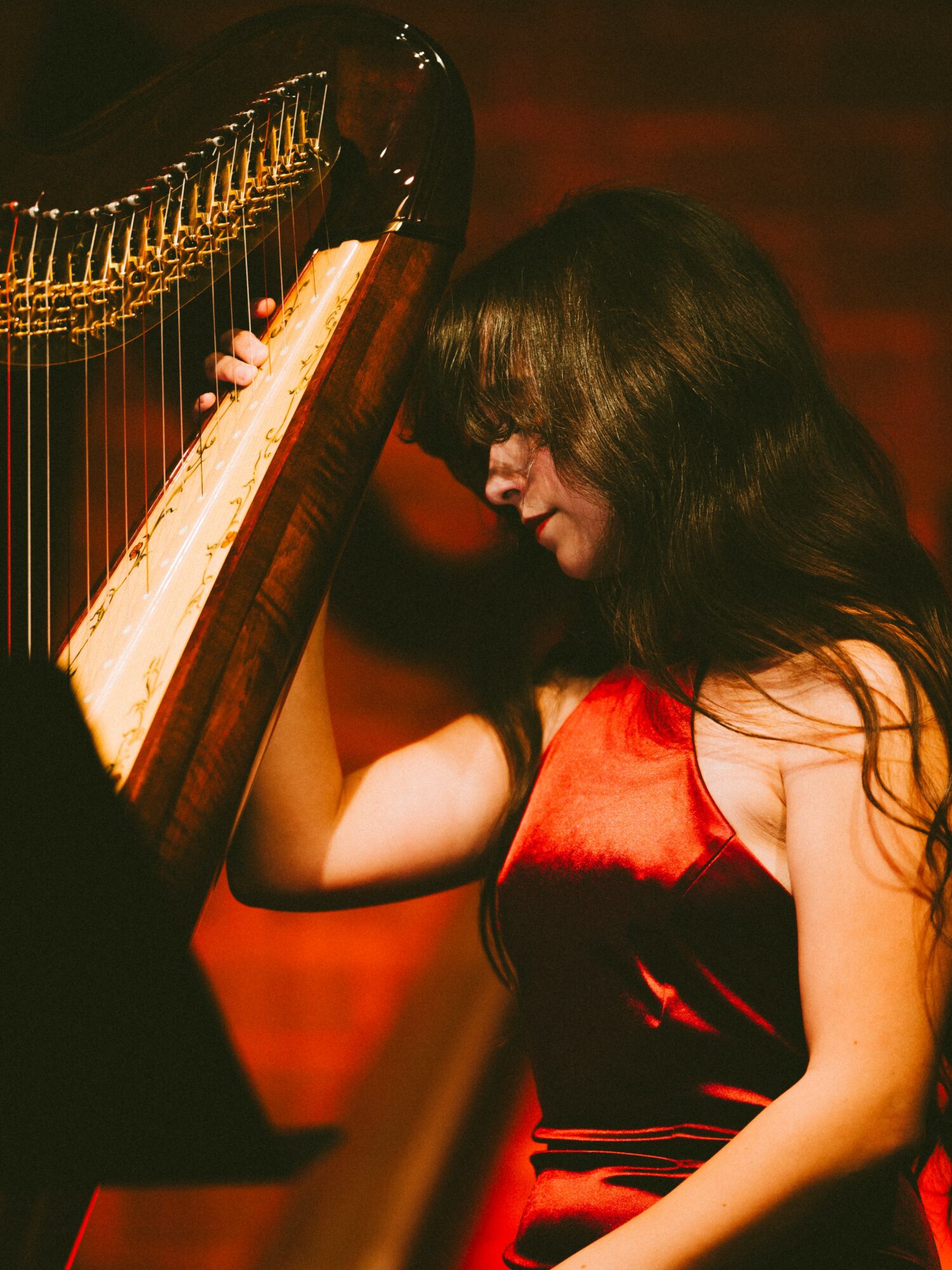 Woman with long dark hair playing a grand piano, wearing a red dress, in a dimly lit room.