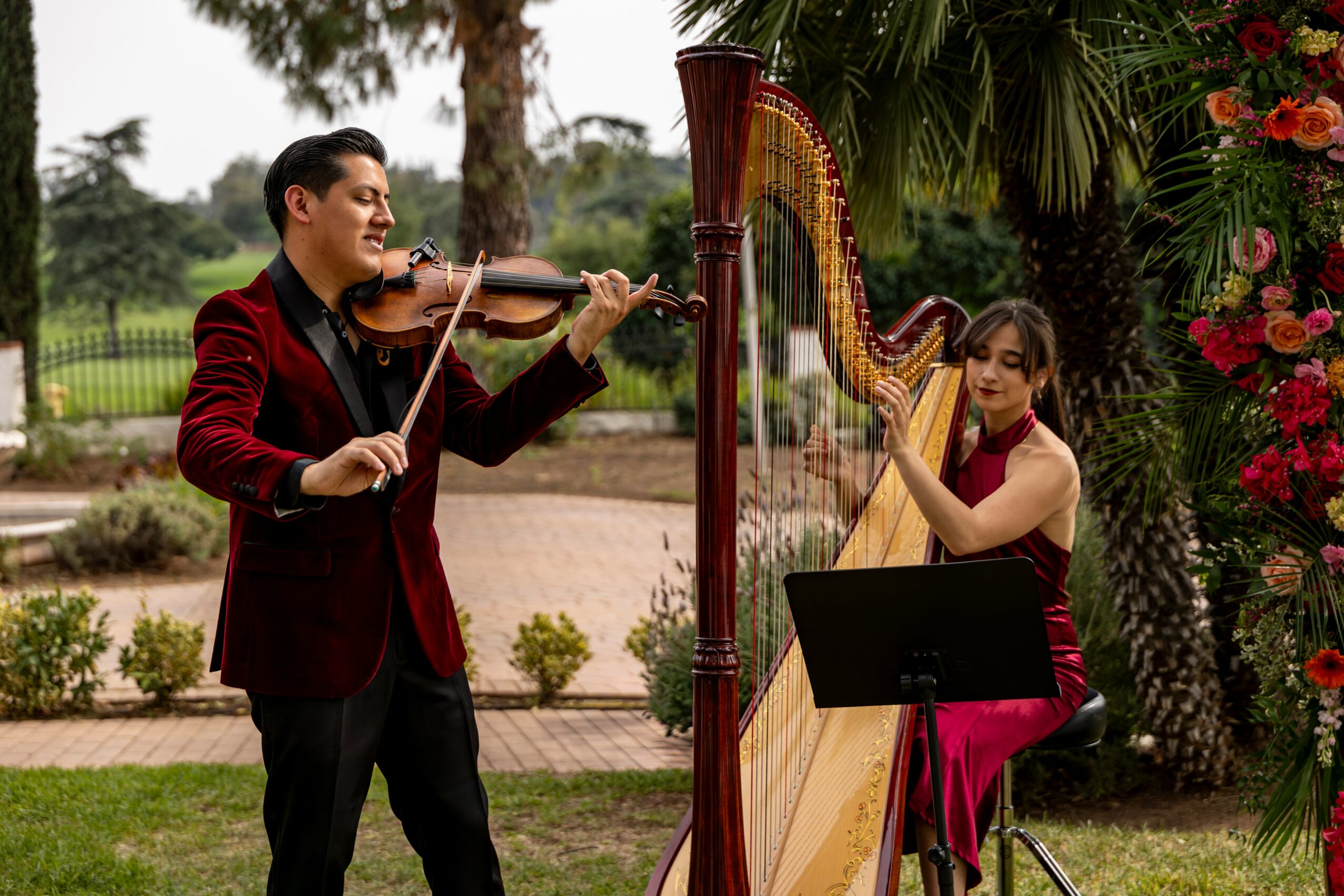 Two musicians, a man playing violin and a woman playing harp, outdoors with trees and flowers.