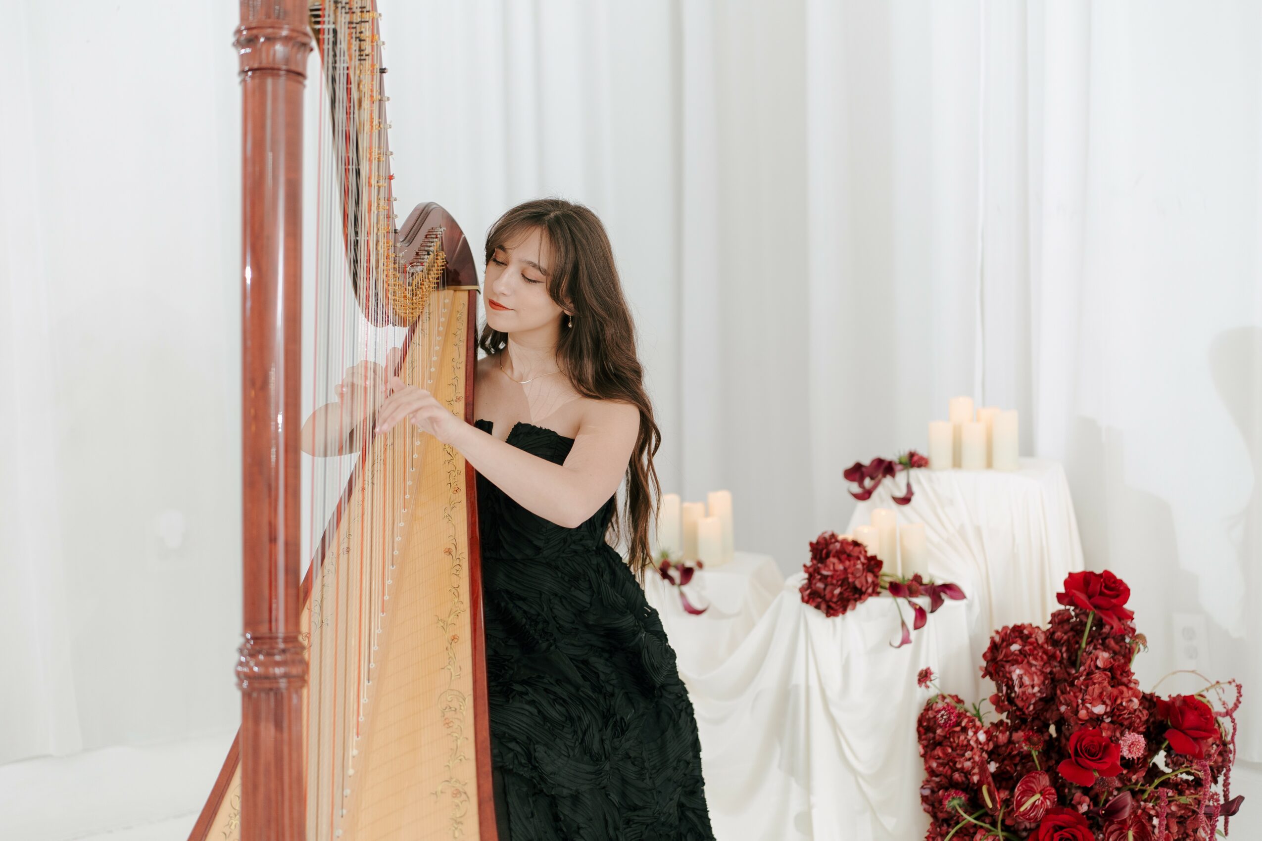 Young woman in black dress playing harp in a bright room with white curtains and red flowers.
