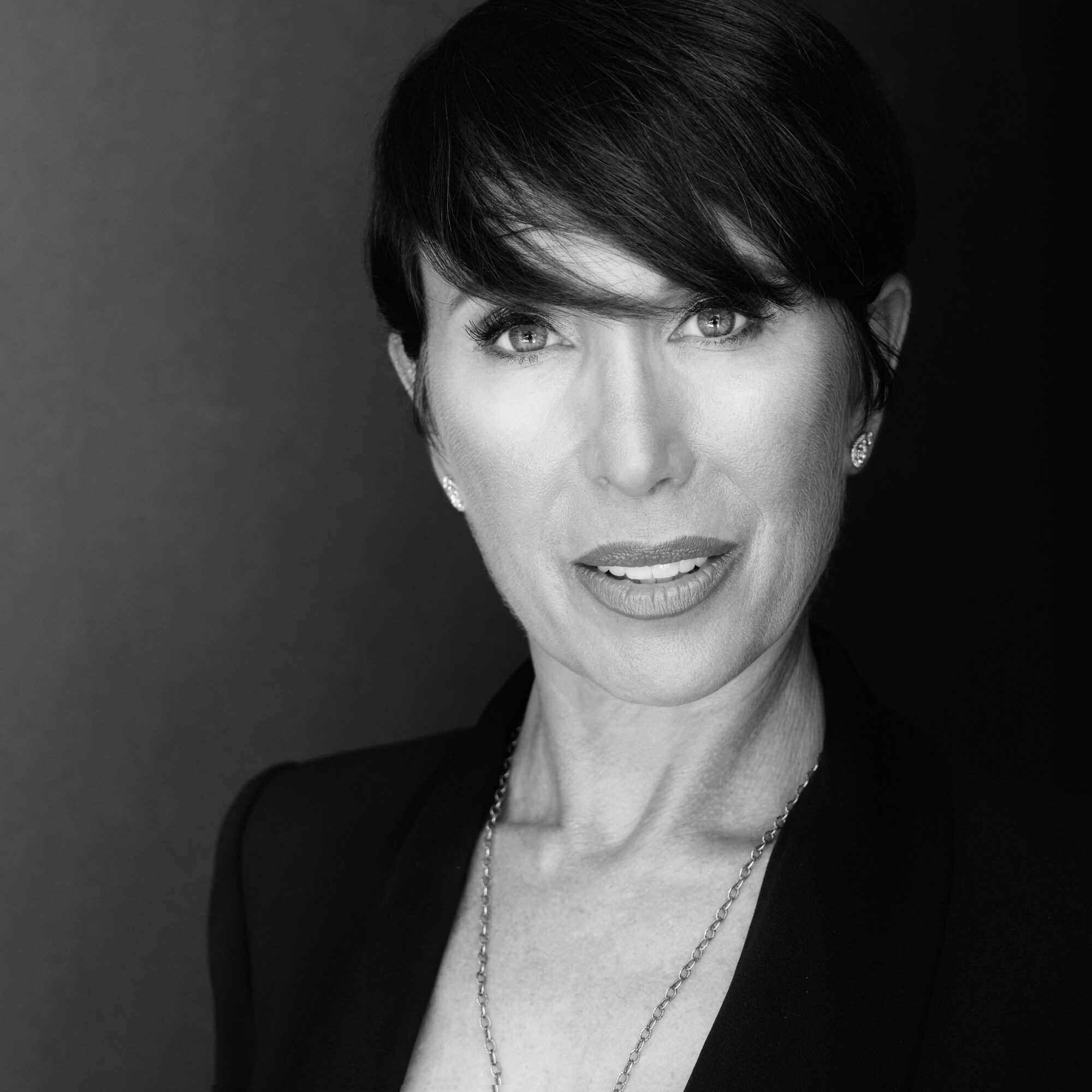 Woman with short dark hair, wearing a blazer and necklace, looking at camera.