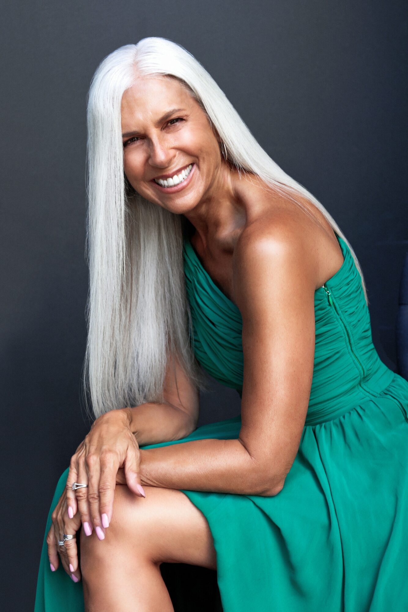 Smiling woman with long white hair wearing a teal dress, sitting and leaning forward.
