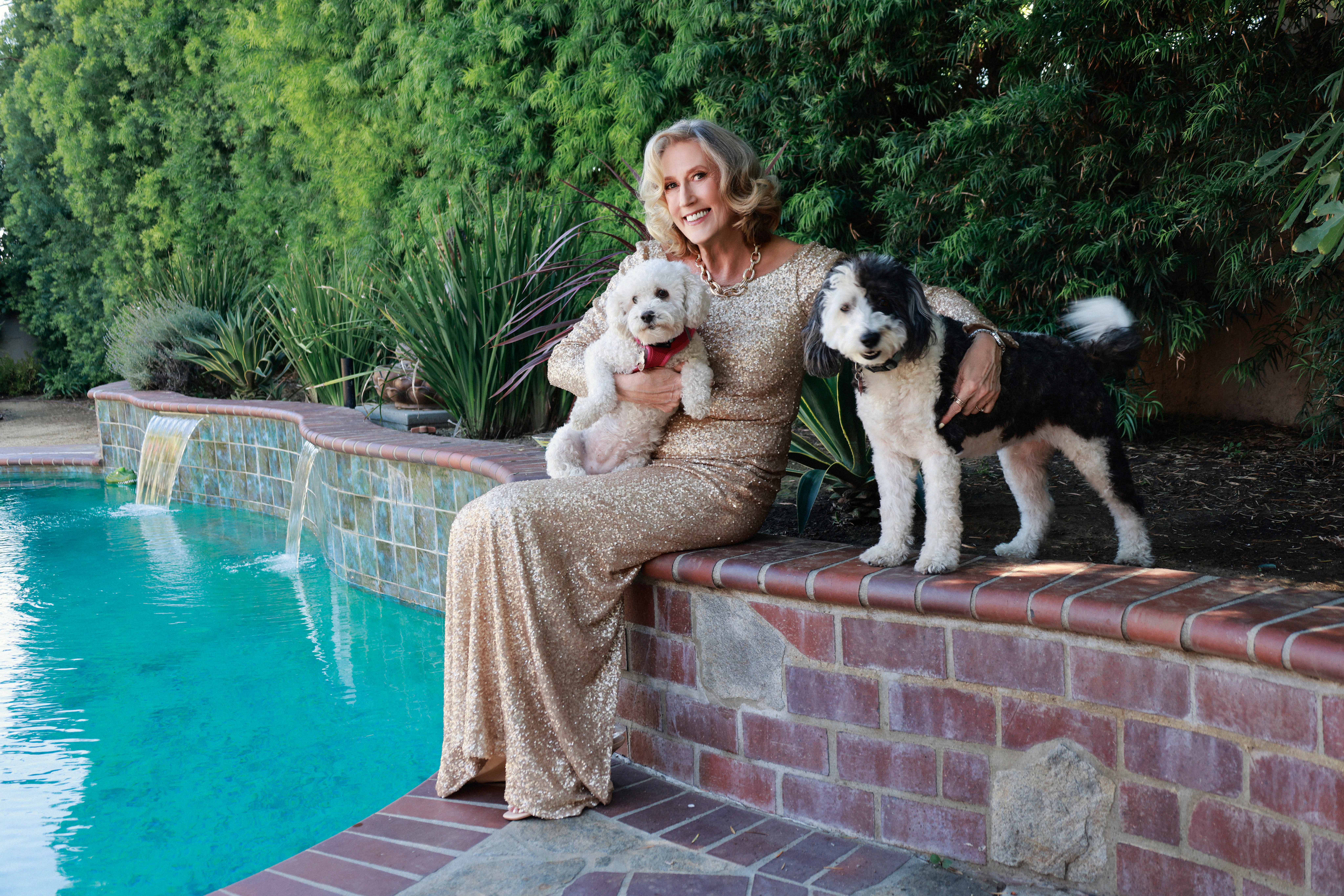 Woman sitting by pool with two dogs, one in her arms and one beside her, surrounded by greenery.