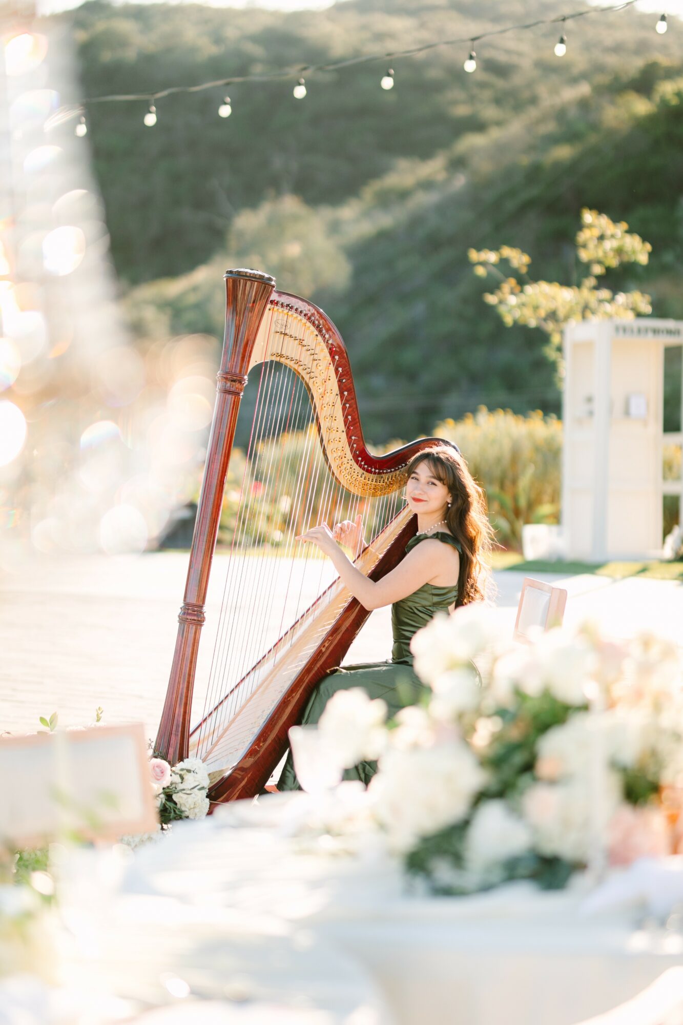 Young woman playing a harp outdoors with trees and string lights in the background.