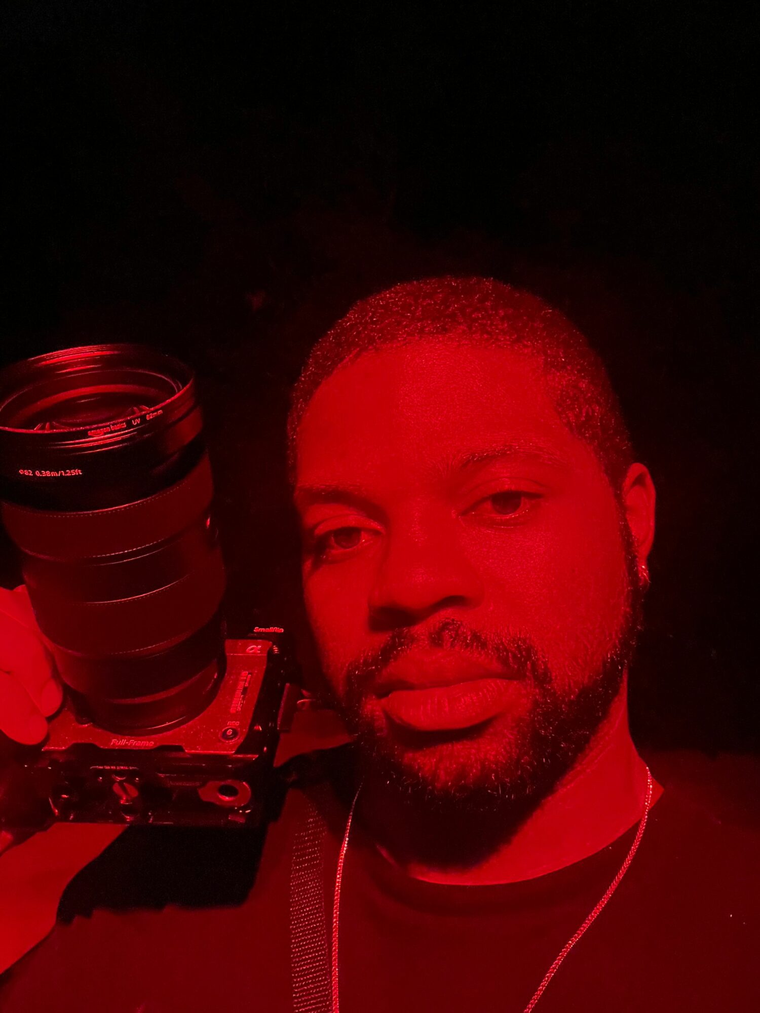 Person with short hair and beard, illuminated by red light, holding a camera, in a dark environment.