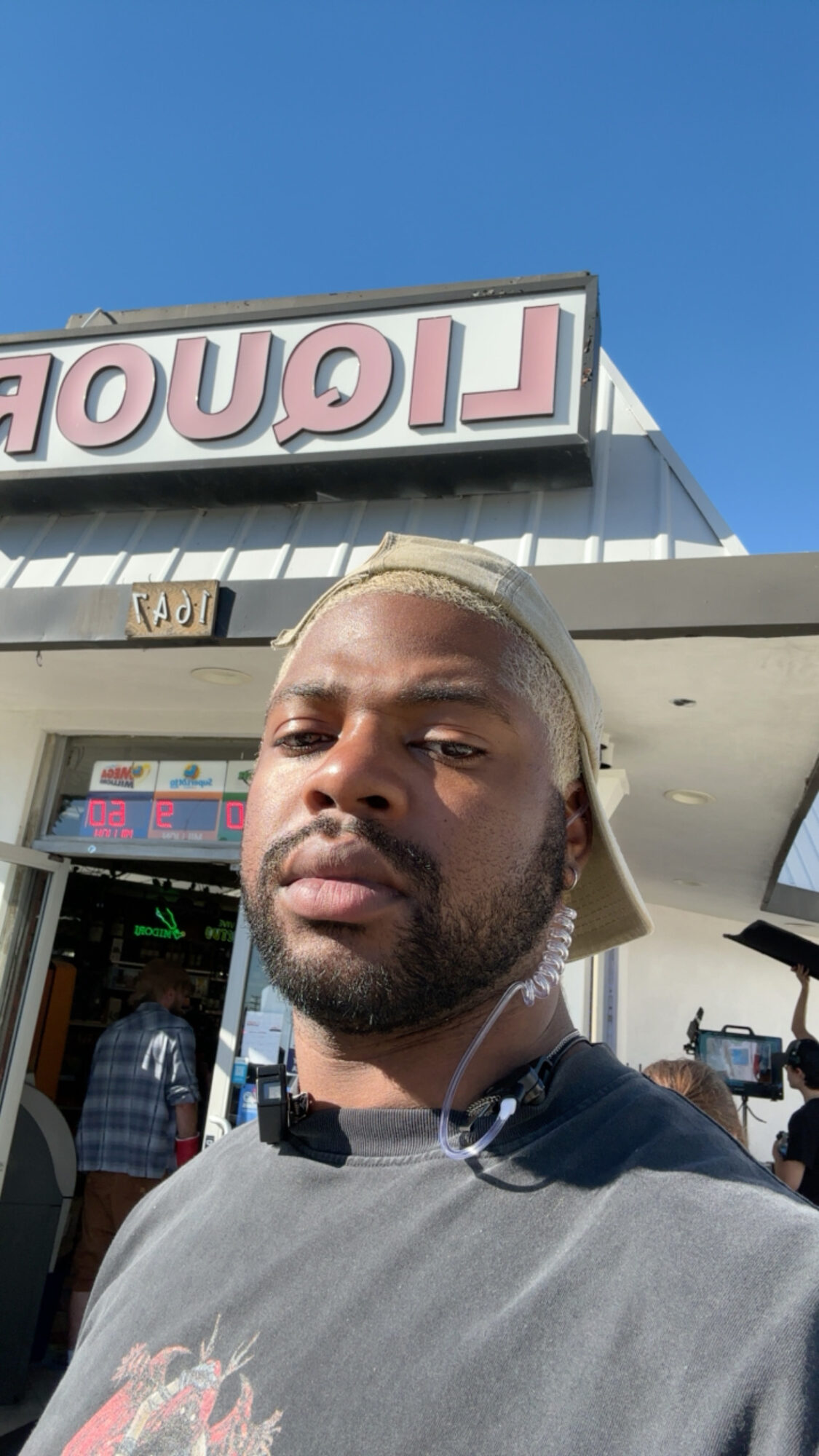 Young man with a beard and blonde hair, wearing a gray shirt, standing outside a store with a sign and blue sky.