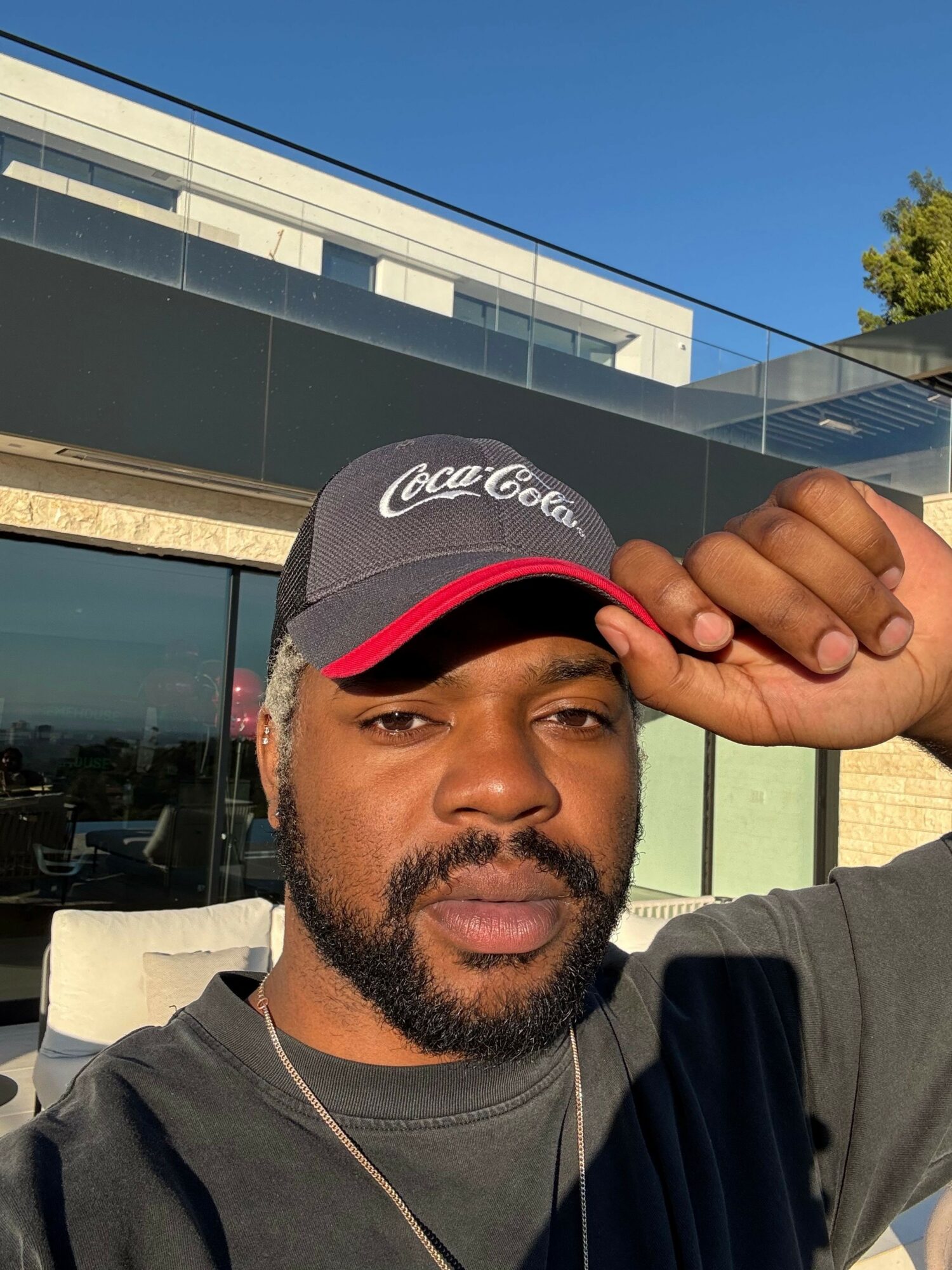 Man wearing a black Coca-Cola cap, adjusting it outdoors with modern building and blue sky in background.
