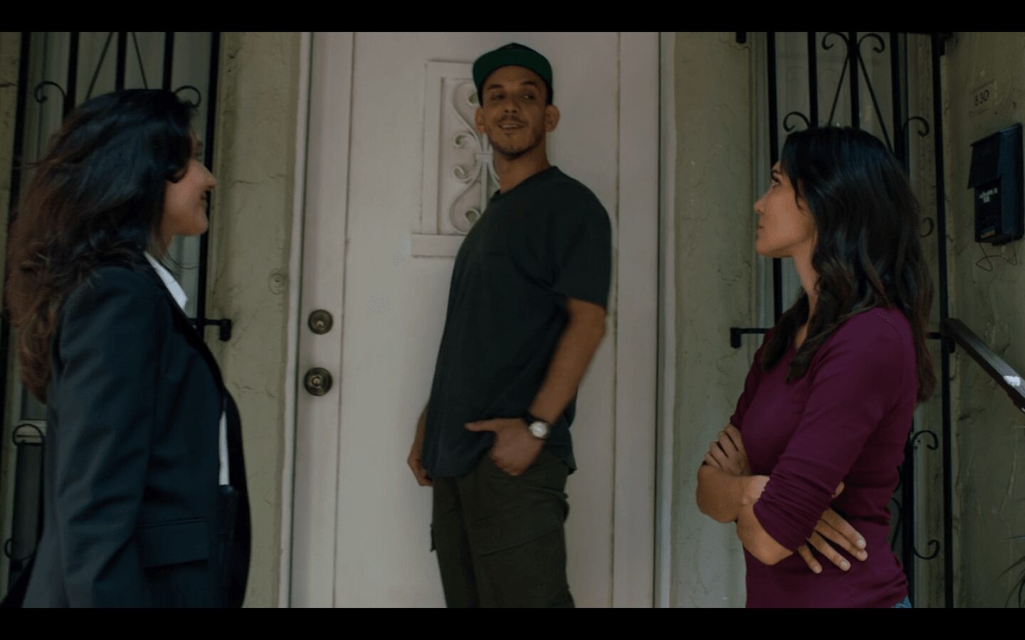 Three people standing outside a door, engaged in conversation, with a man in the center wearing a cap and casual clothes.