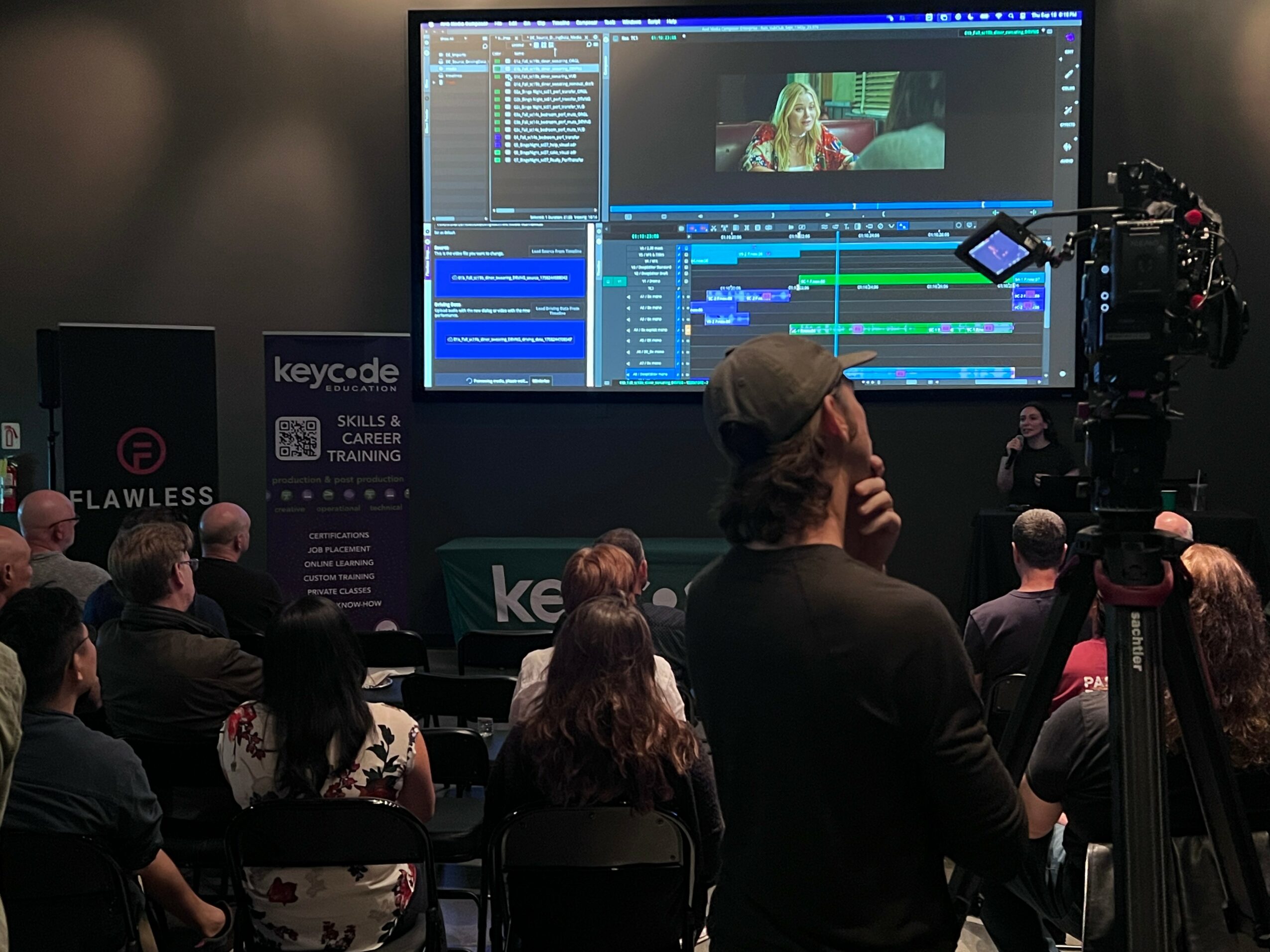 Audience watching a presentation with a large screen showing video editing software and a woman on screen.