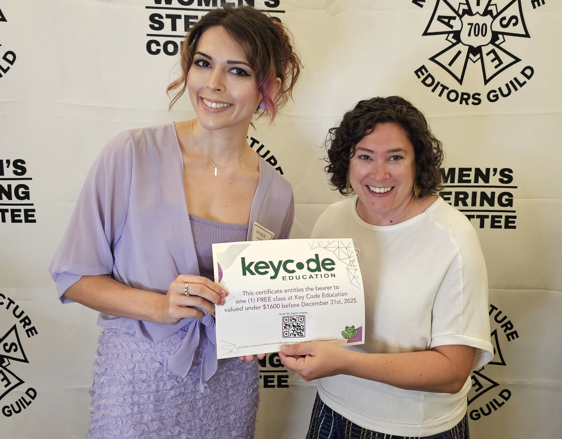 Two women holding a certificate, smiling, standing in front of a backdrop with text and logos.