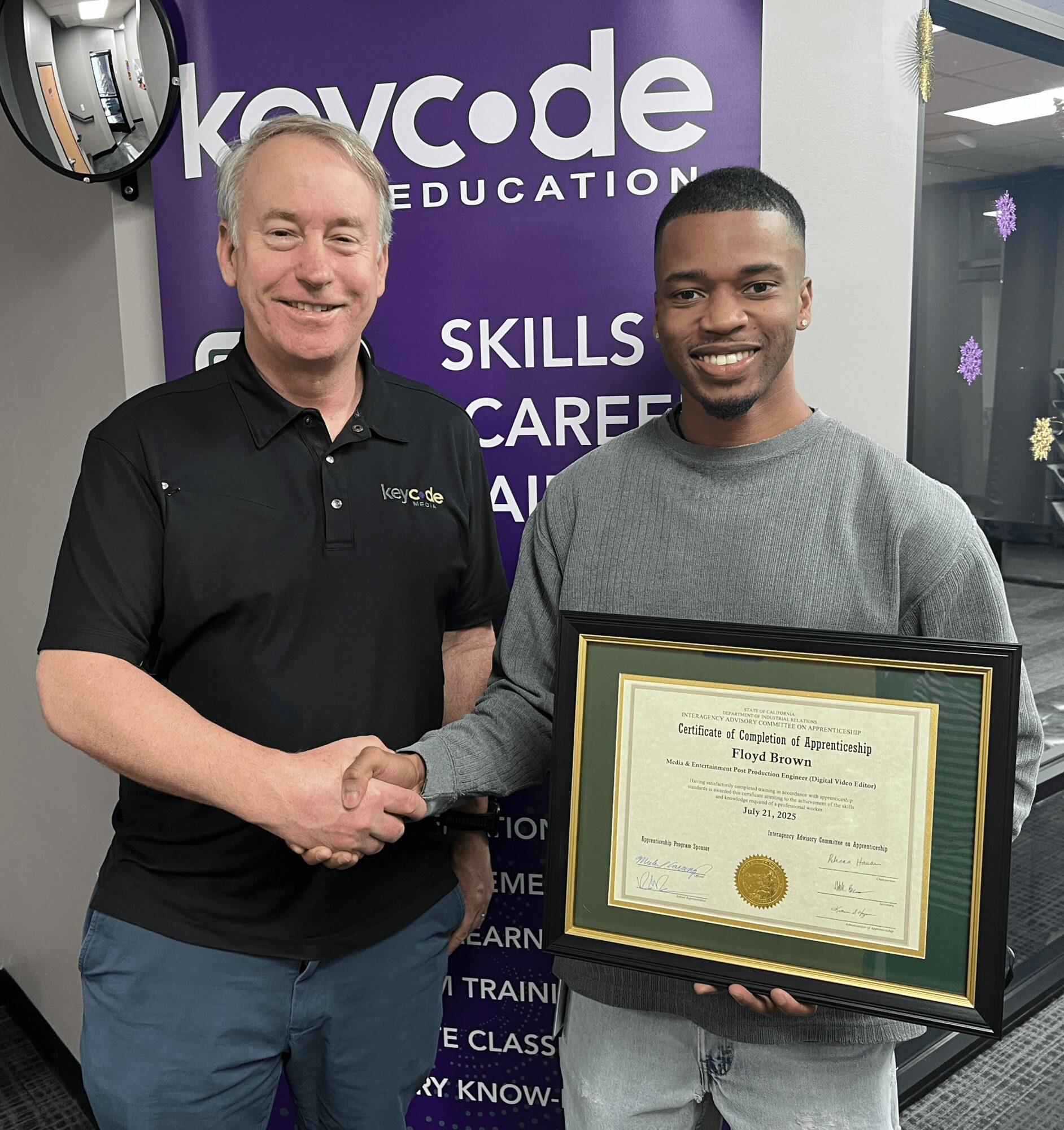 Two men shaking hands, one holding a framed certificate, standing in front of a purple and white banner.