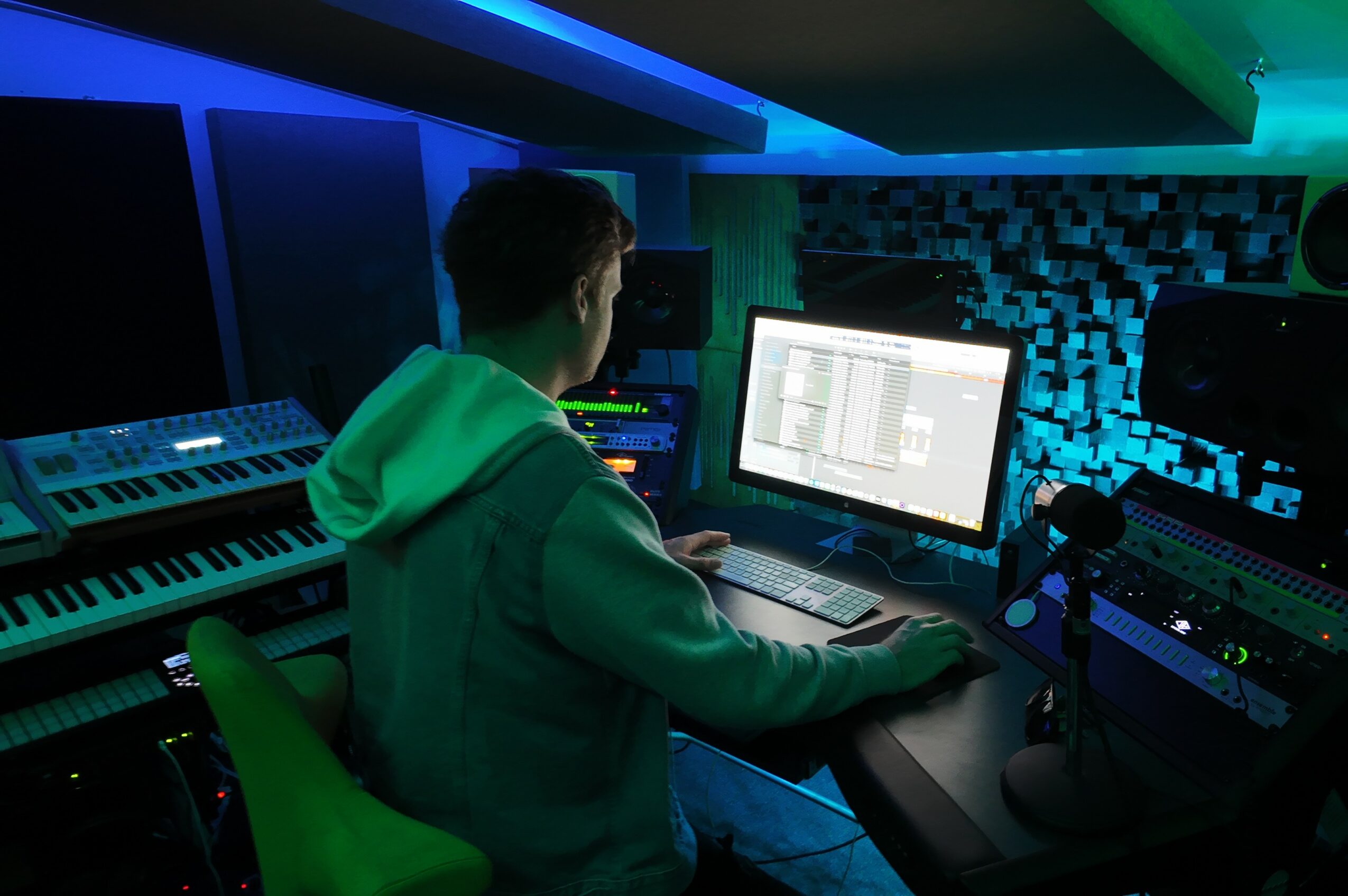 Person working at a computer in a music studio with keyboards and audio equipment, illuminated by blue and green lights.