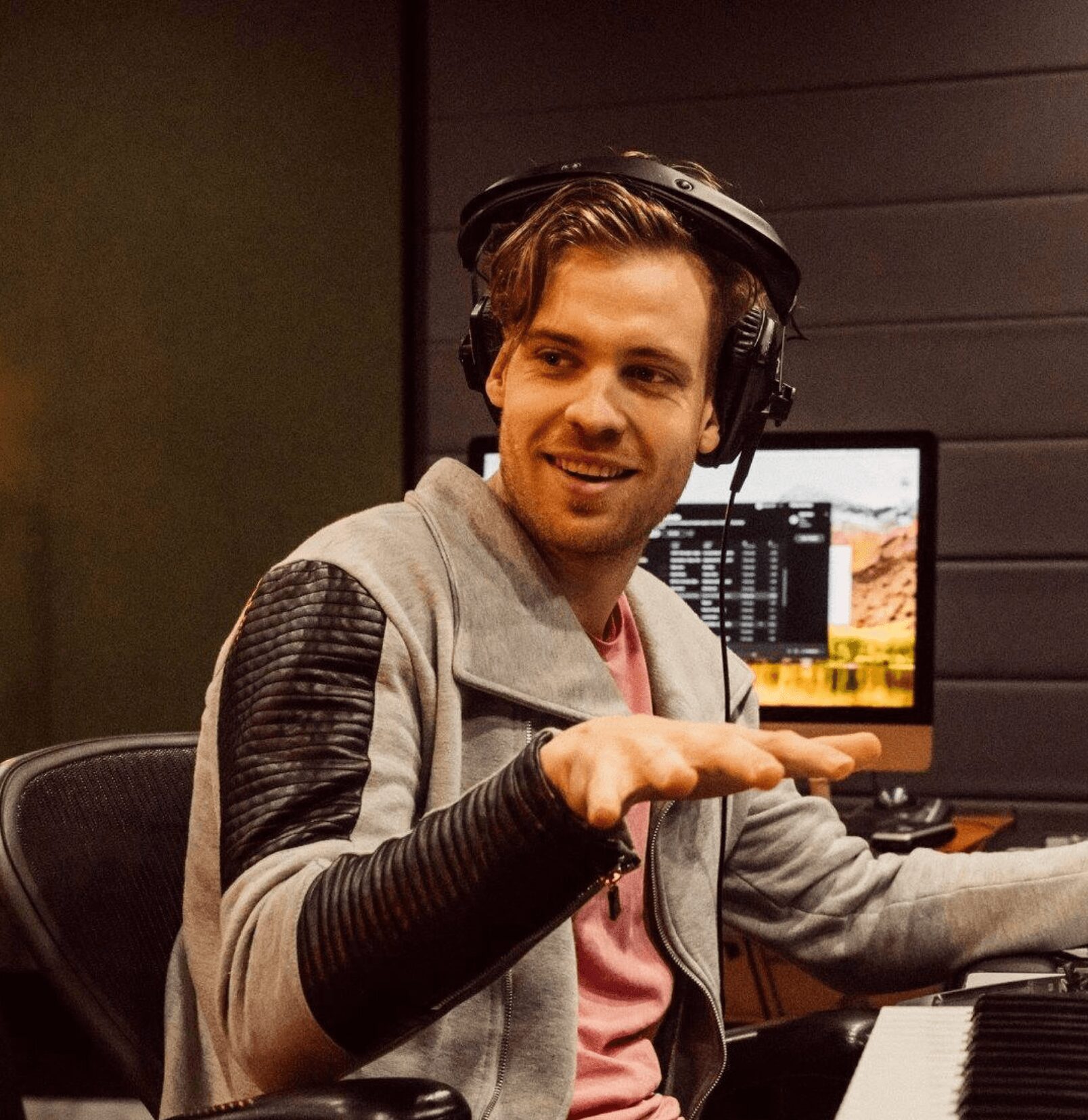 Young man wearing headphones, smiling, gesturing with hand, sitting at a keyboard in a recording studio.