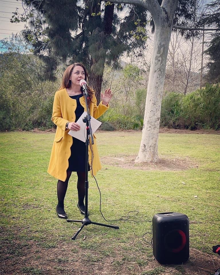 Woman speaking into microphone outdoors, holding papers, wearing yellow coat and black dress, with speaker nearby.