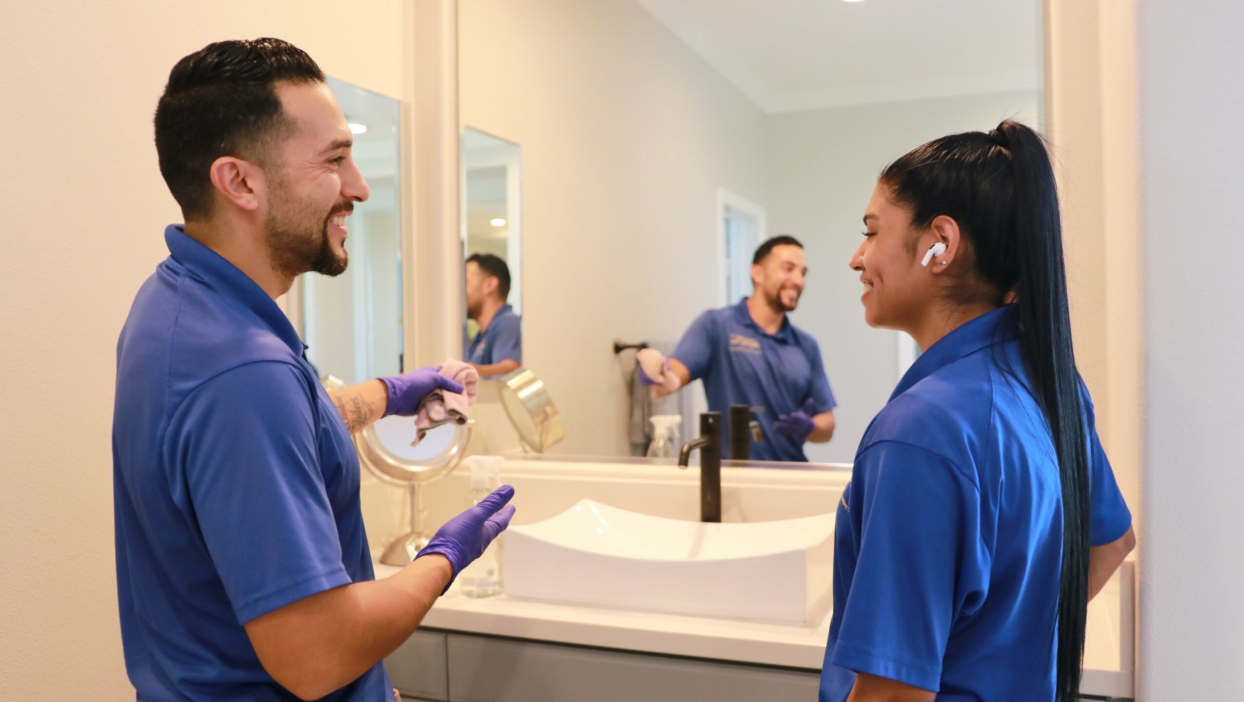 Two people in blue uniforms talking in a bathroom with a mirror and sink, smiling at each other.