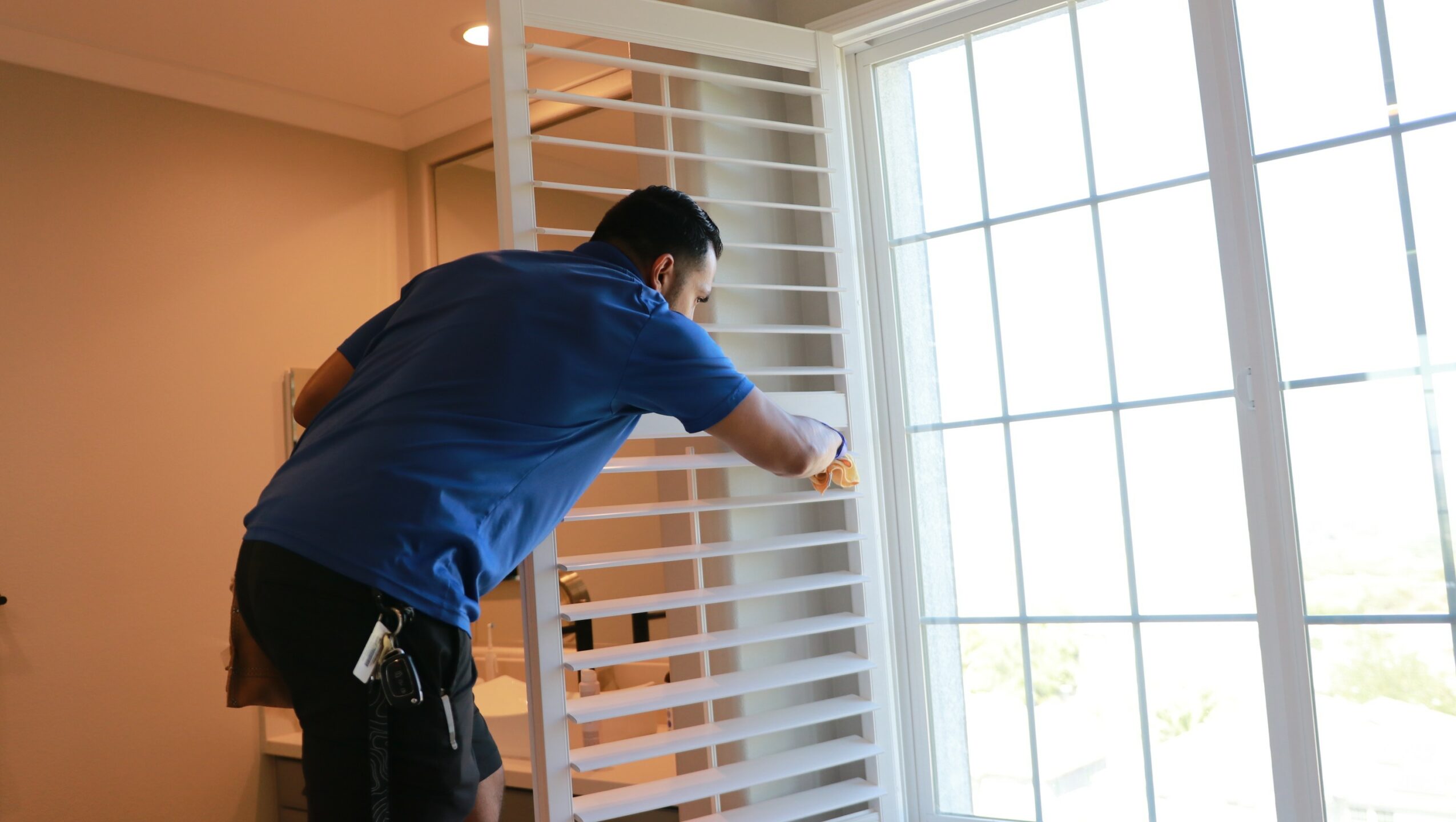 Person cleaning window blinds near large window with sunlight streaming in.