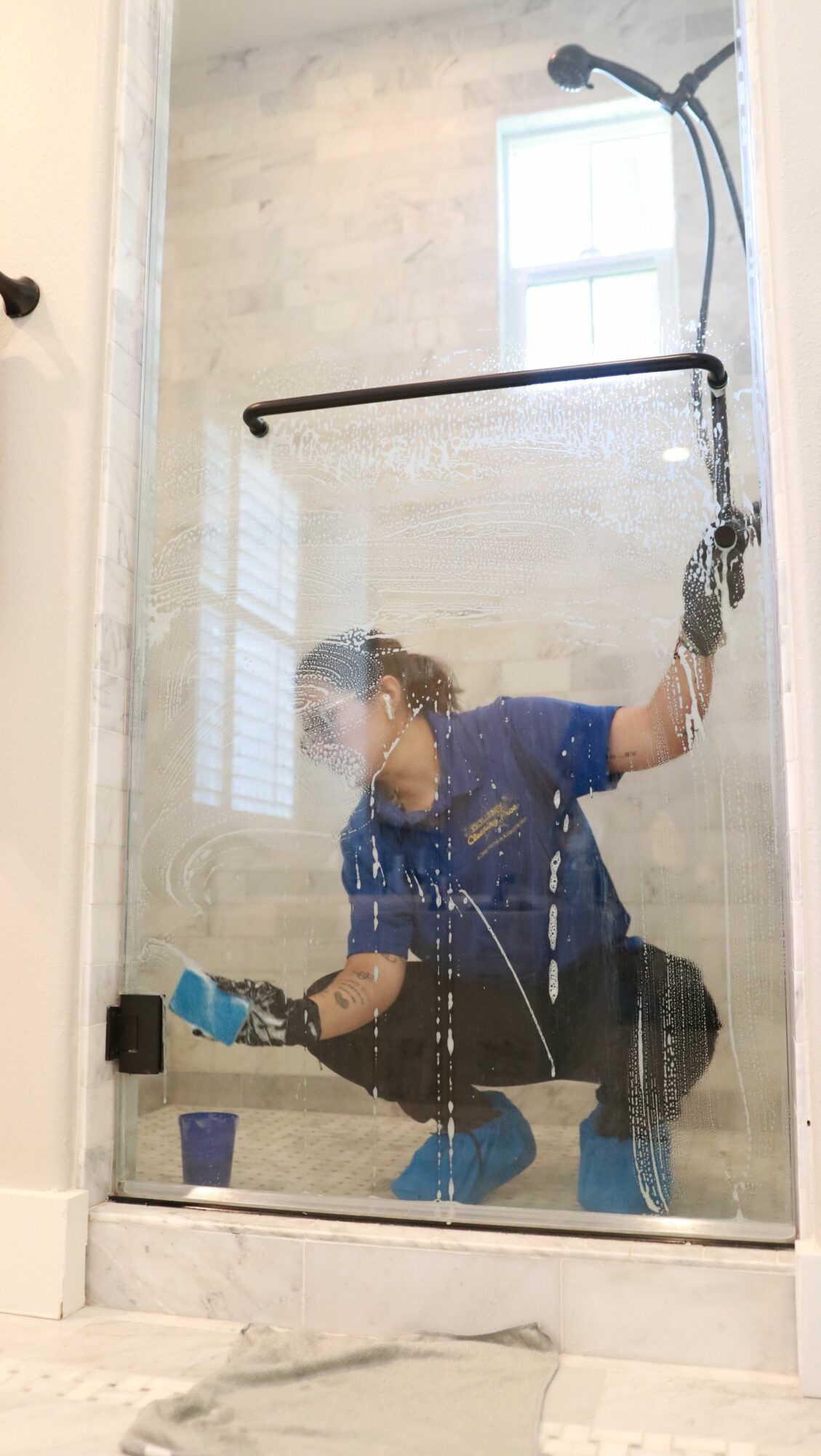 Person cleaning glass shower door with a cloth and spray, wearing gloves and a blue shirt.