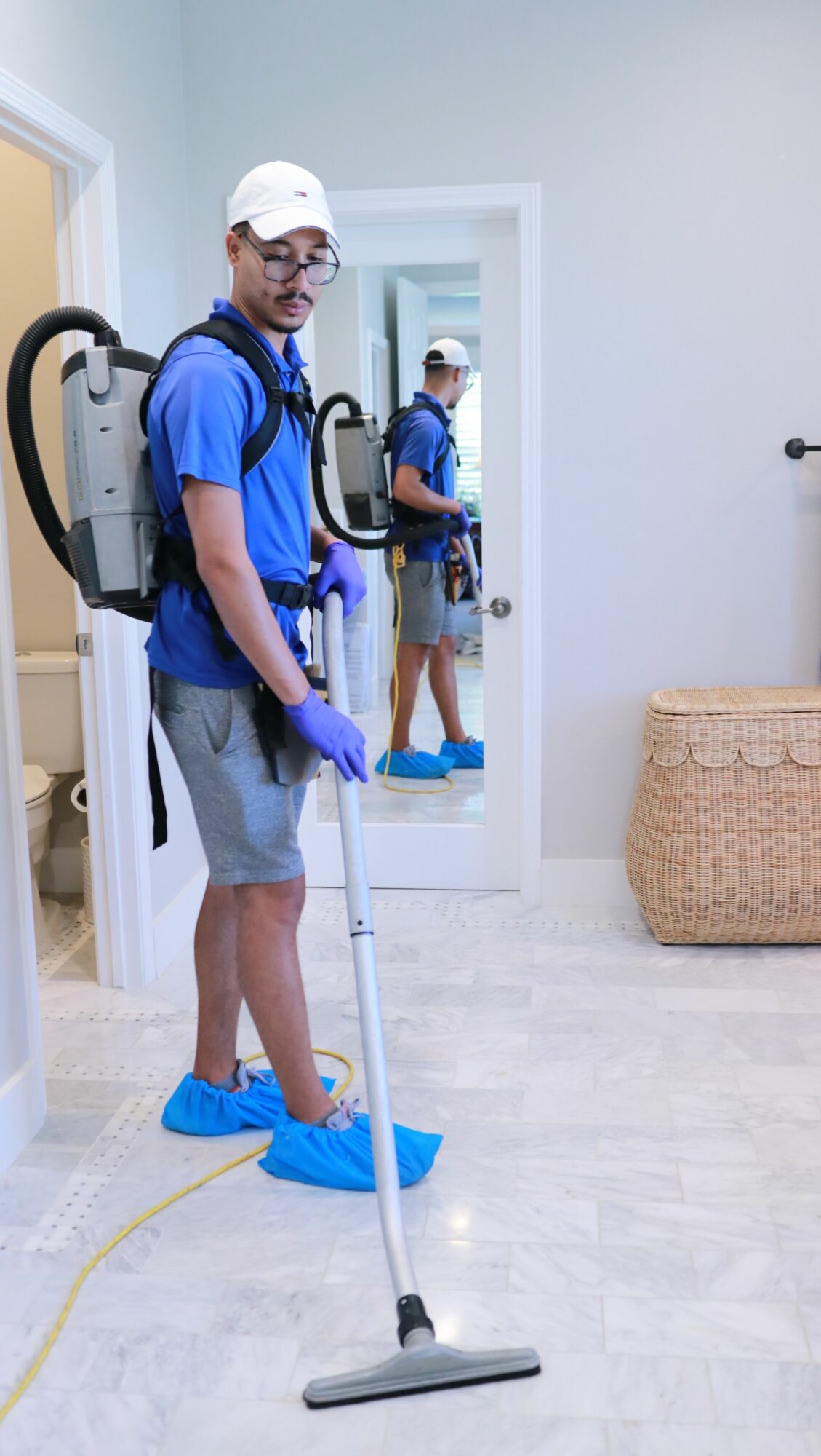 Person cleaning floor with vacuum, reflected in mirror, wearing blue shoe covers, gloves, and casual clothing.