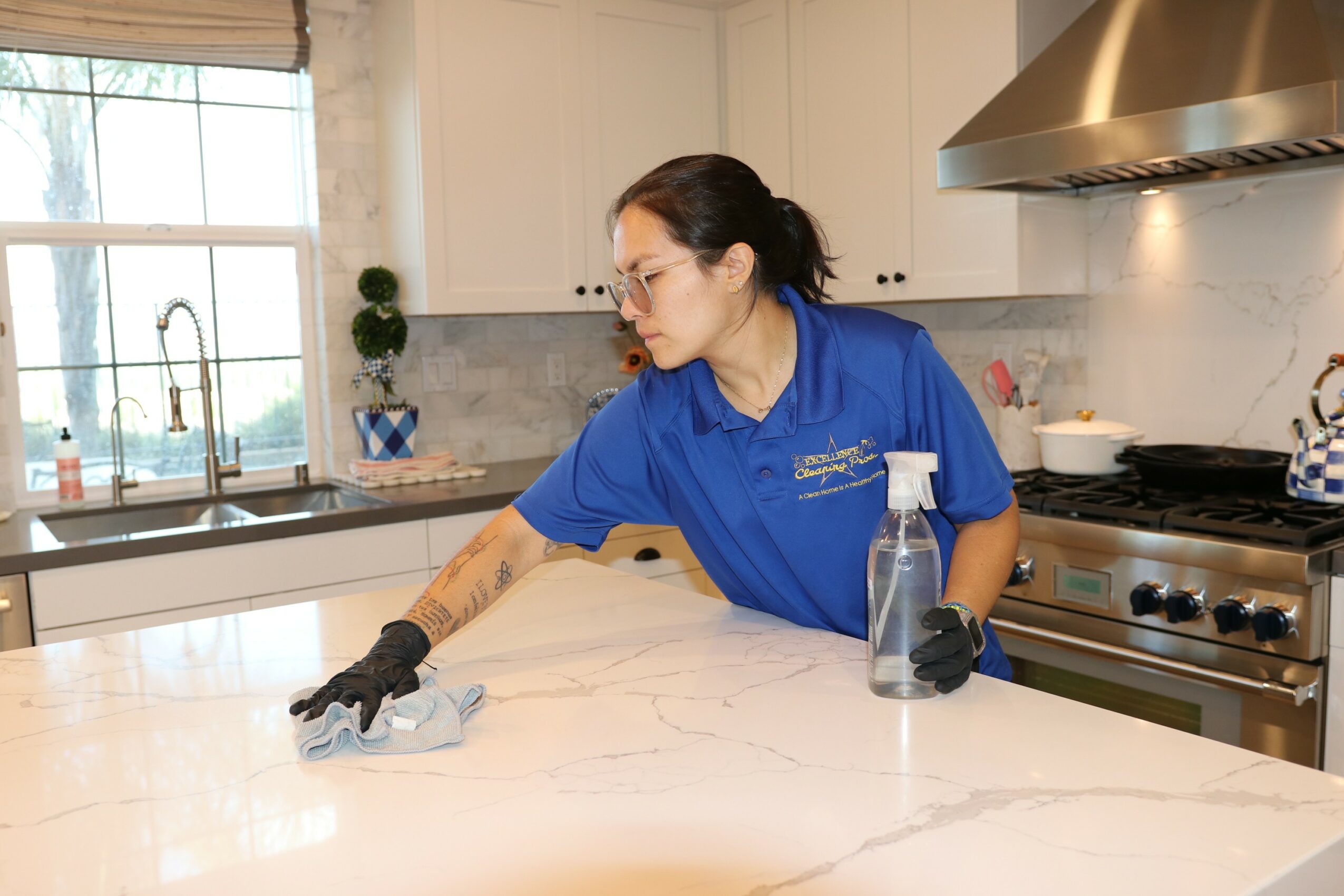 Woman in blue uniform cleaning a kitchen countertop with a cloth, spray bottle nearby, in a bright kitchen.