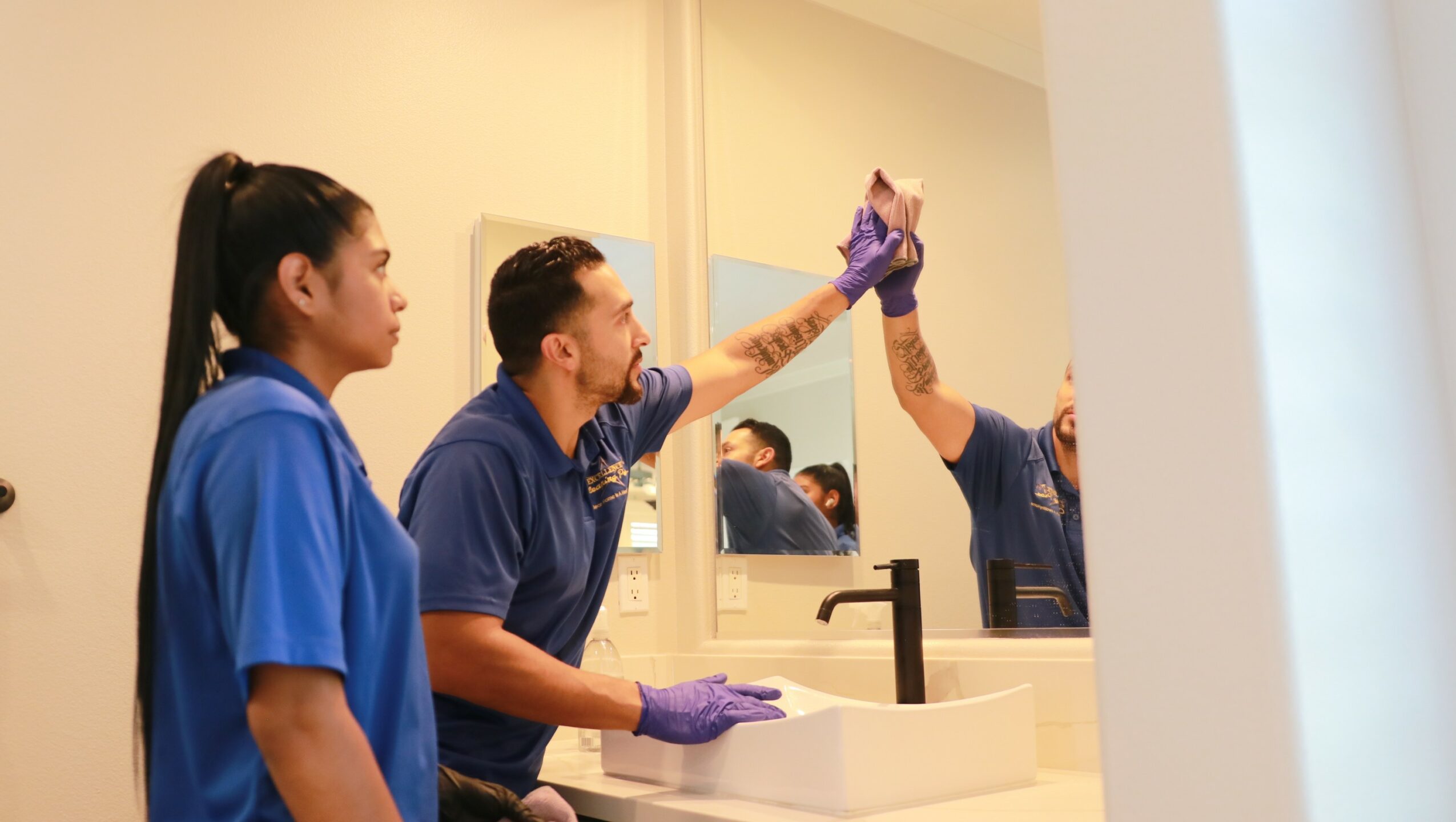 Three people in blue uniforms in a bathroom, one cleaning a mirror with a cloth, others observing.