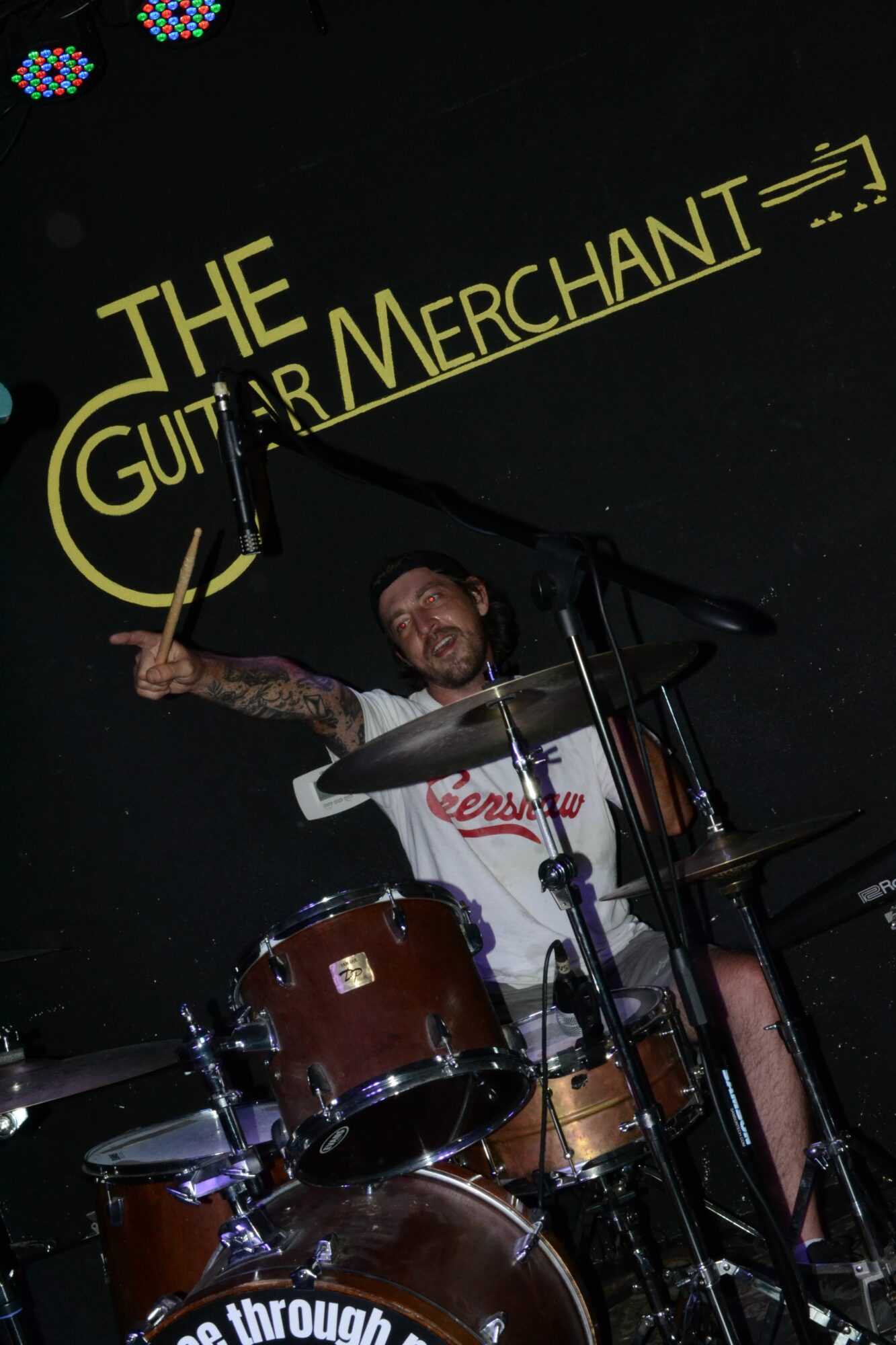 Person playing drums on stage with a sign reading 'The Guitar Merchant' behind them.