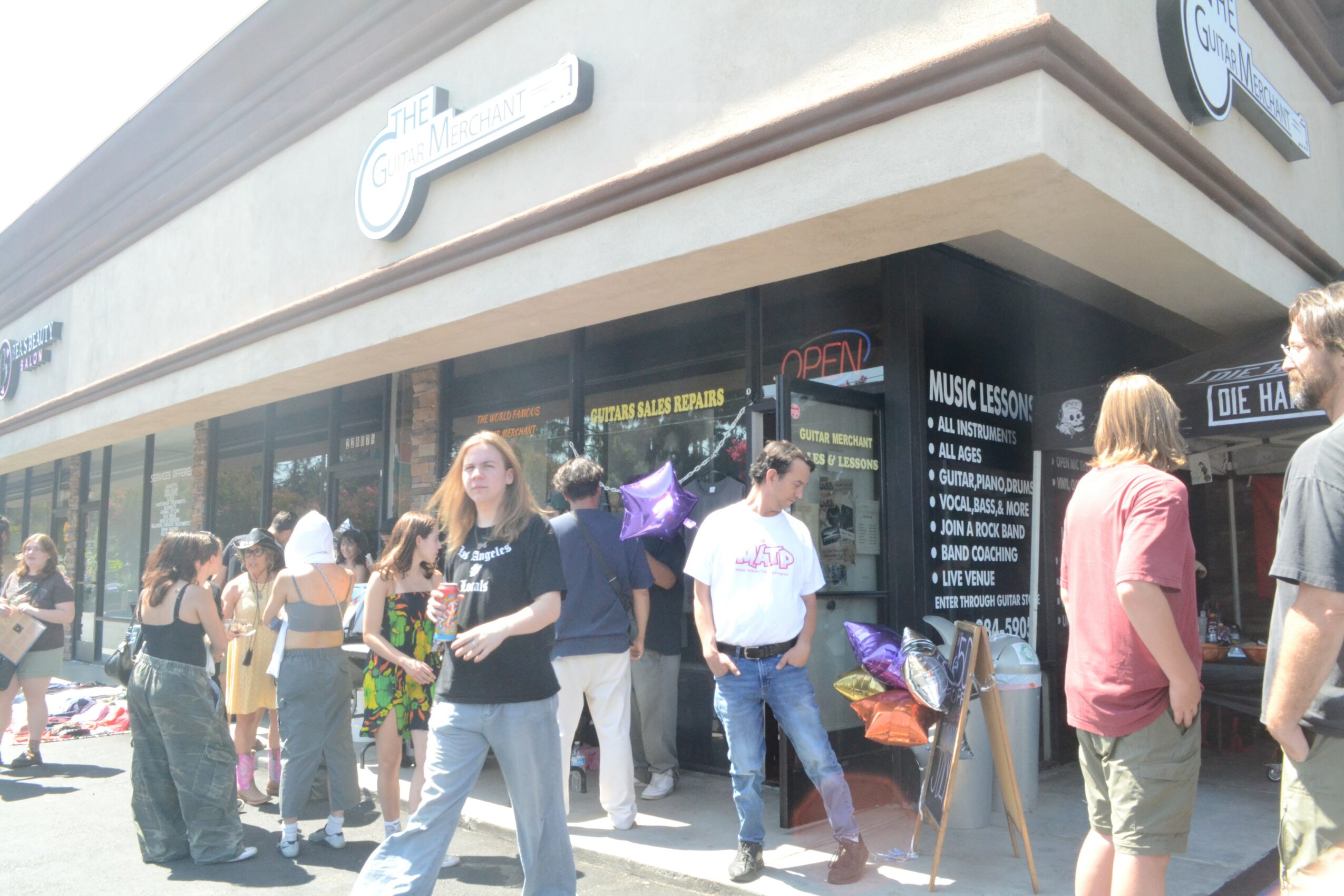 People gather outside a store with large glass windows and a signboard, some holding balloons and bags.