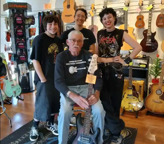 Four people in a music store with guitars on the wall, some standing and one sitting with a guitar.