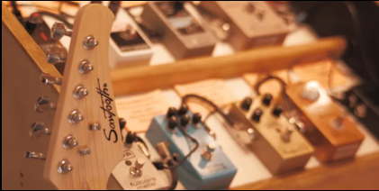 Close-up of guitar headstock with tuning pegs and part of a wooden neck, on a cluttered surface.