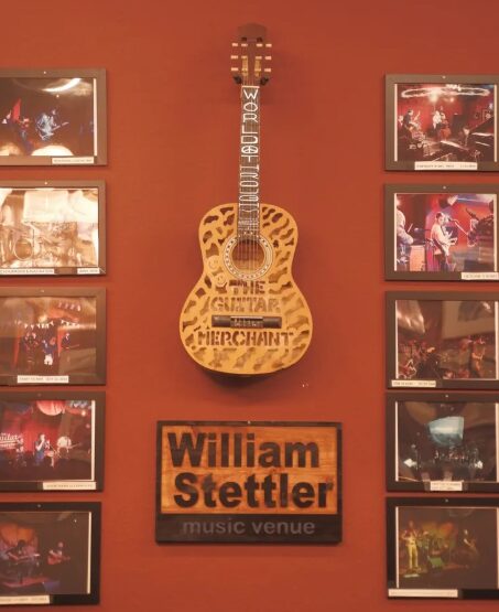 Guitar mounted on a wall with framed pictures on both sides, and a sign below reading 'William Stettler music venue'.