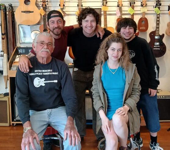 Group of five people, three men and two women, in a music store with guitars on the wall behind them.