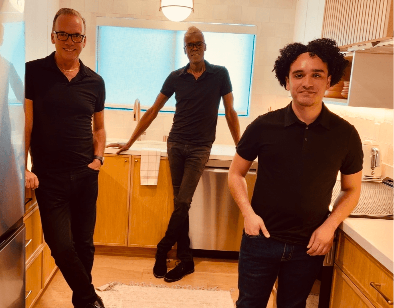 Three men standing in a kitchen, two in front and one in the background, with a window and kitchen appliances.