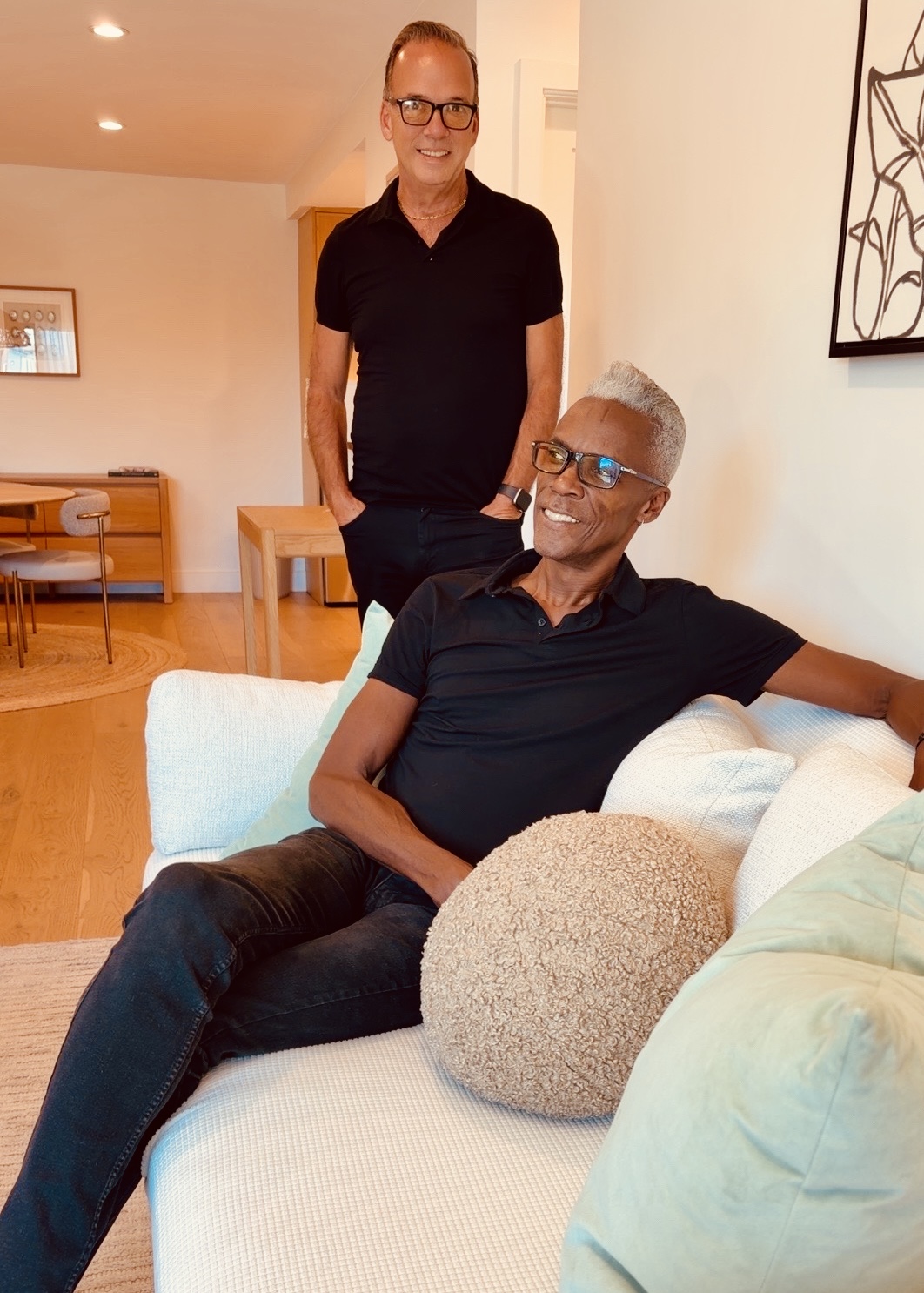 Older man with glasses sitting on a sofa, smiling, with a woman standing behind him in a living room.