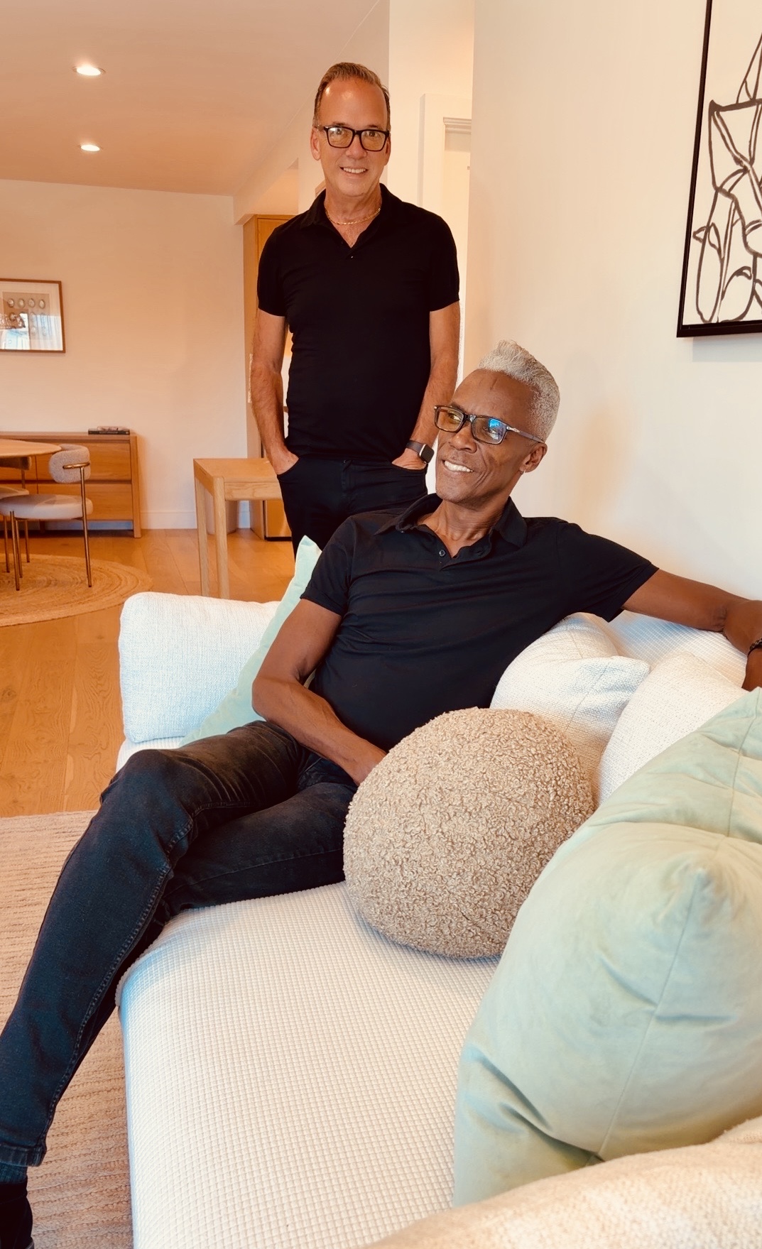 Two people in a living room, one sitting on a sofa and one standing behind, smiling and wearing glasses.