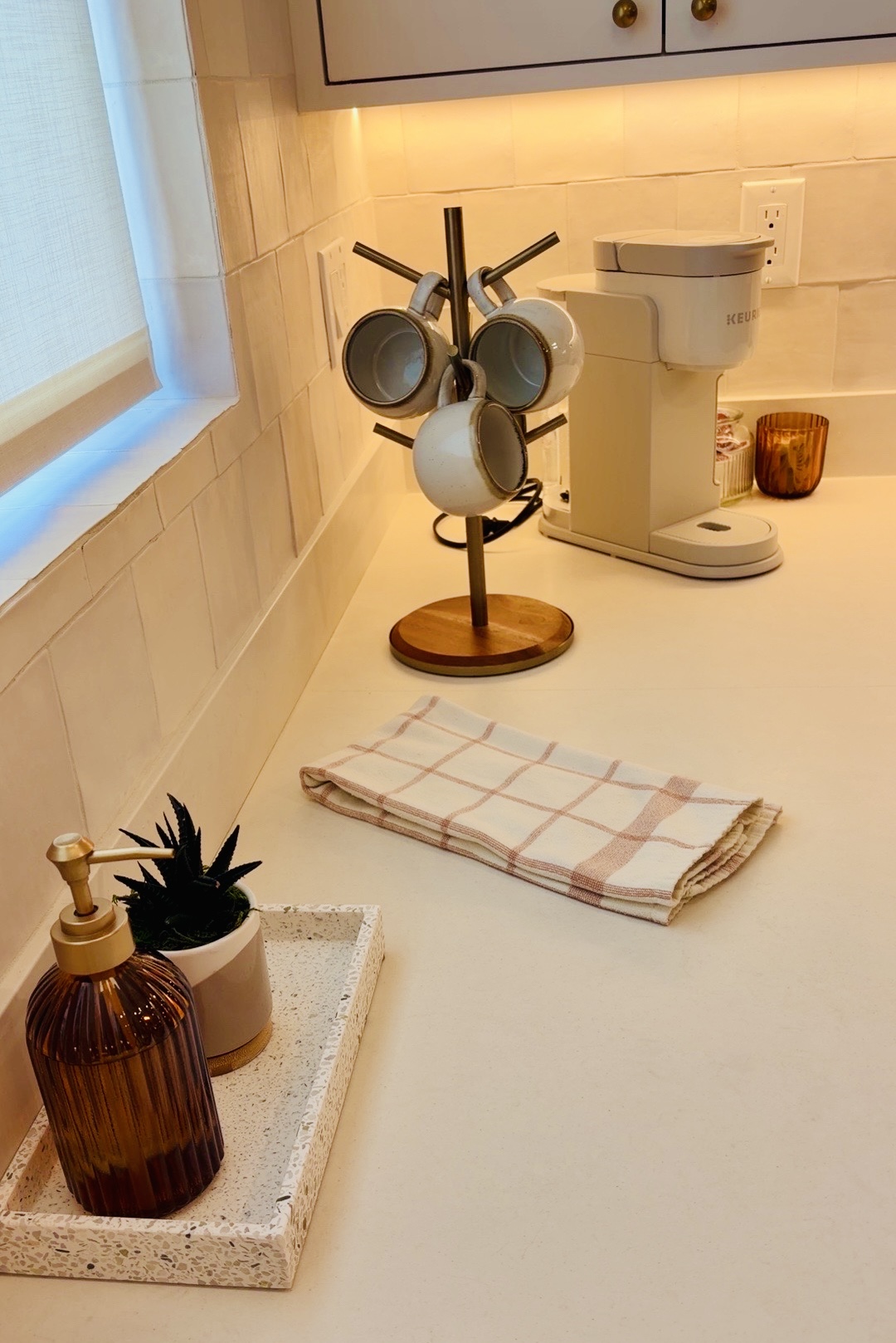 Kitchen countertop with a towel, coffee machine, and a mug holder with cups, near a window and beige tiles.