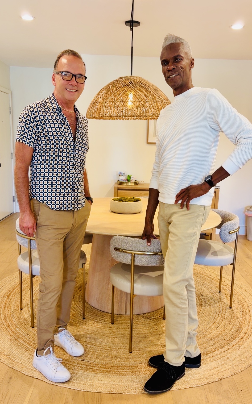 Two men standing in a dining room, smiling, near a round table with chairs, a hanging light, and a beige rug.