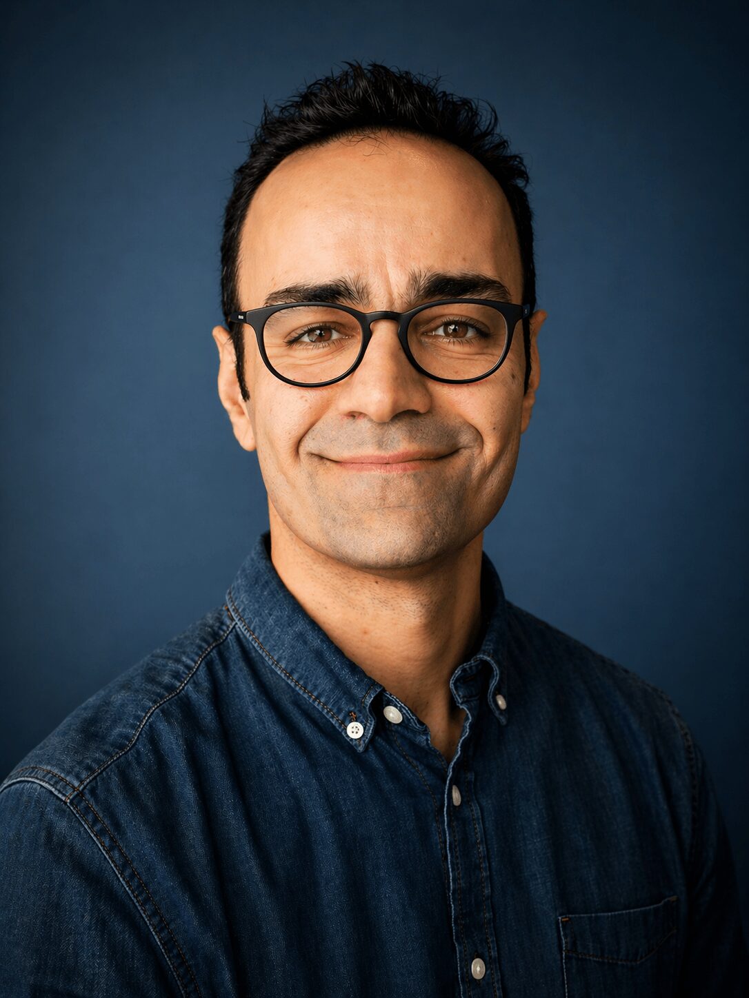 Man with glasses and dark hair, wearing a denim shirt, smiling against a dark blue background.